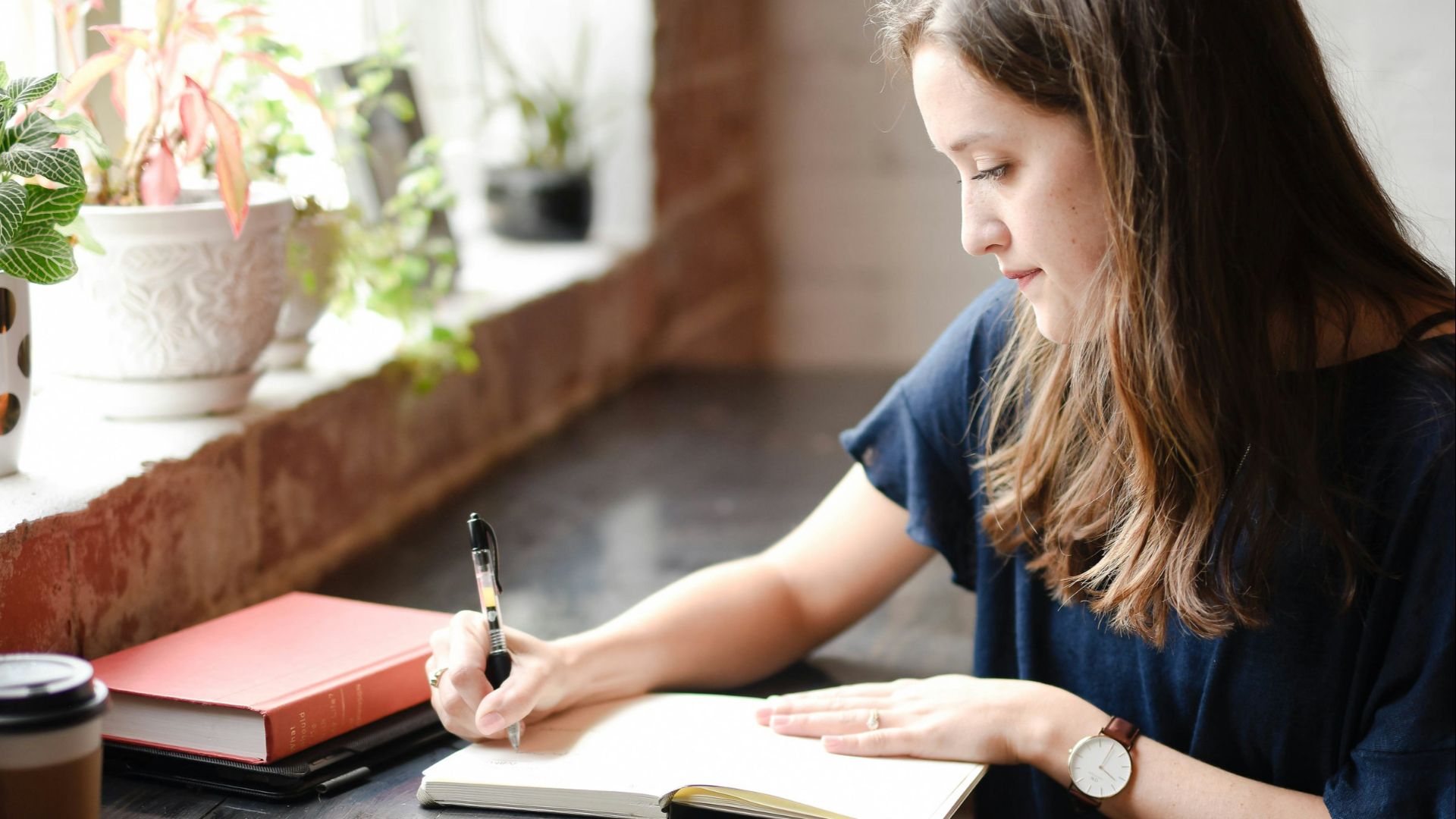 woman sitting in front of black table writing on white book near window