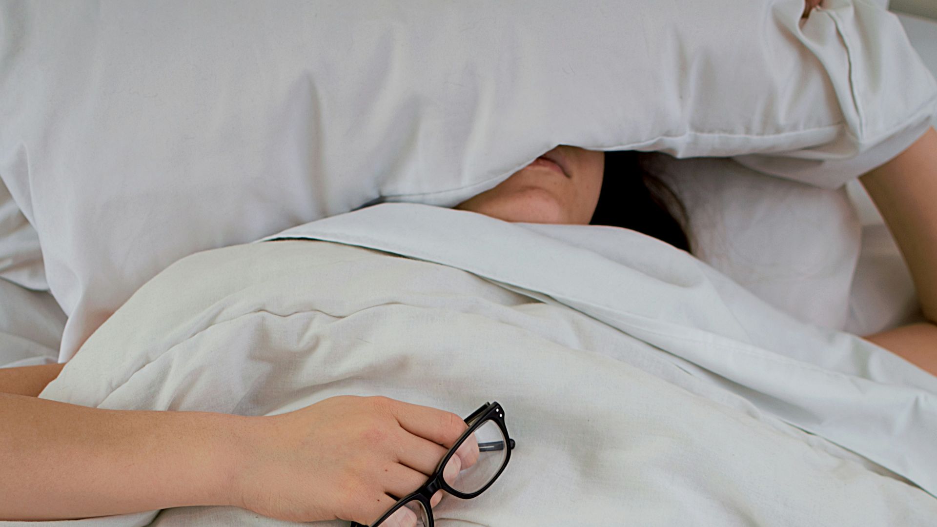 person lying on bed while covering face with pillow and holding eyeglasses