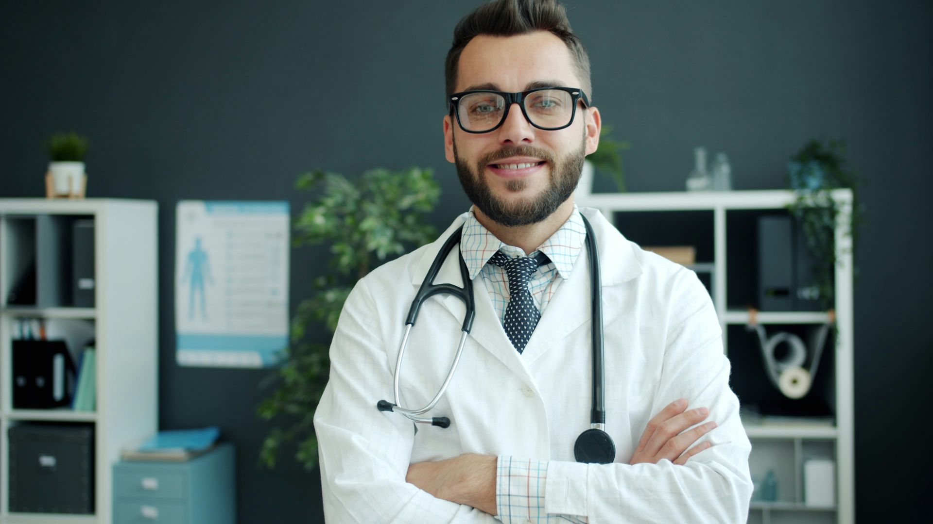 A smiling doctor with glasses and stethoscope stands arms crossed.