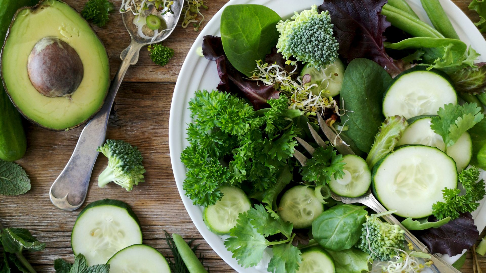 sliced broccoli and cucumber on plate with gray stainless steel fork near green bell pepper, snowpea, and avocado fruit