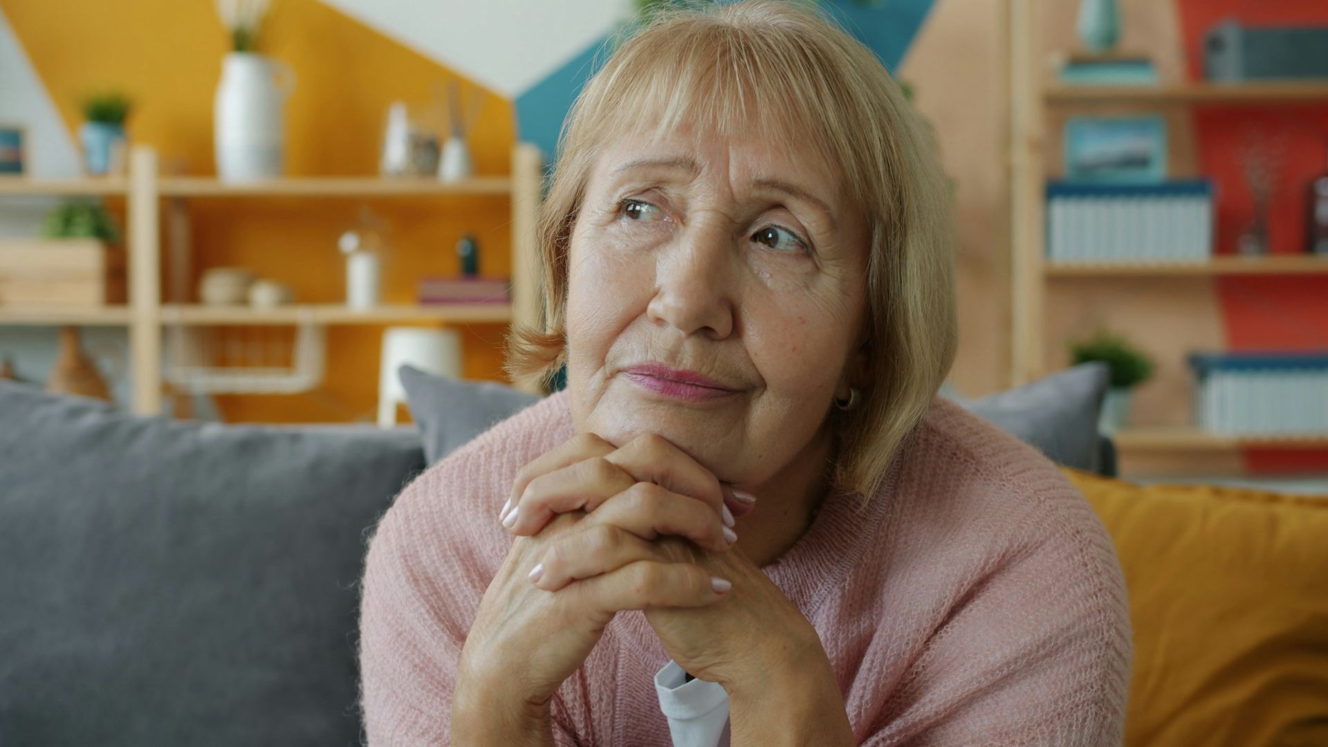 Elderly woman with hands clasped, looking thoughtful.