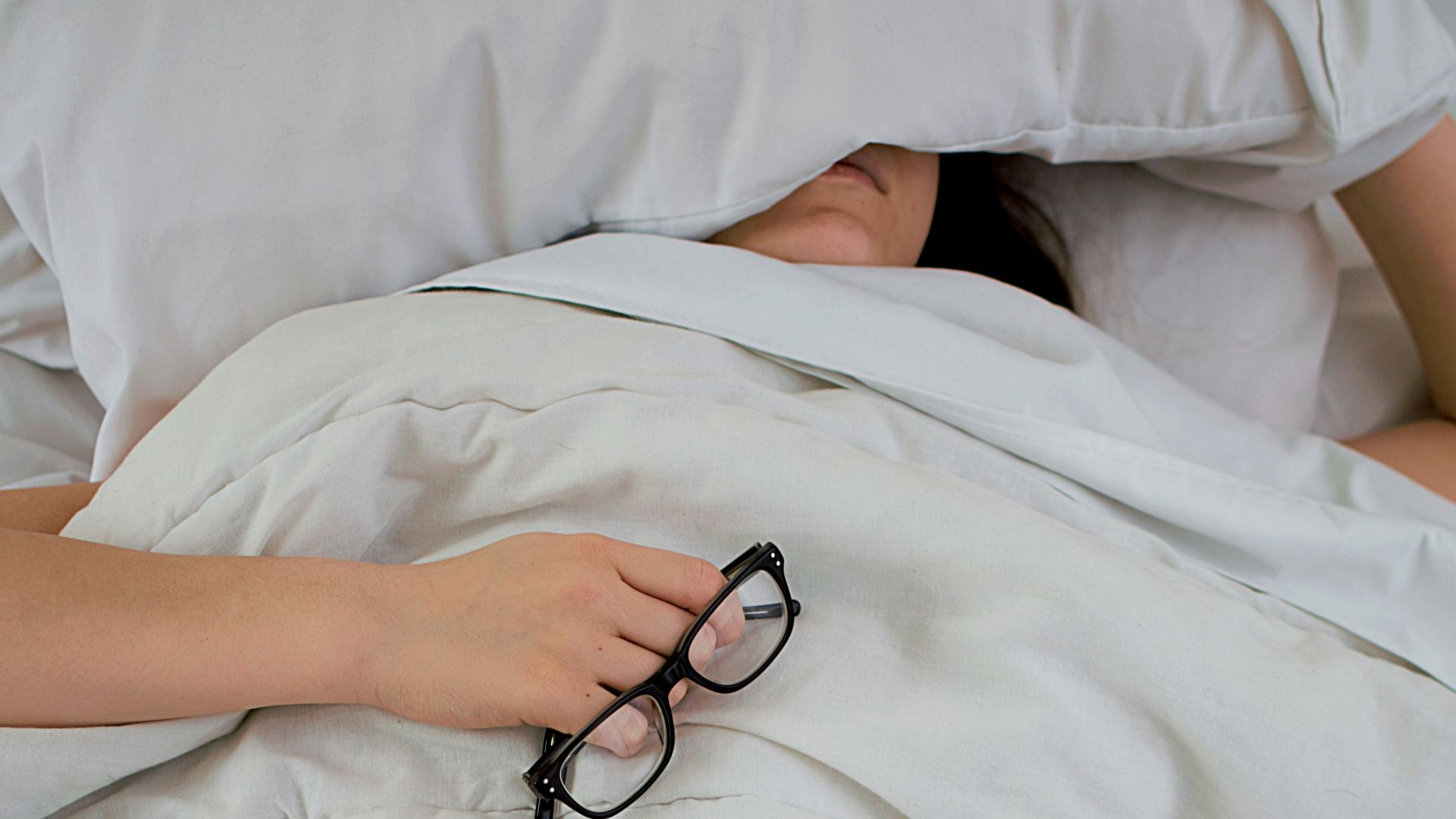 person lying on bed while covering face with pillow and holding eyeglasses