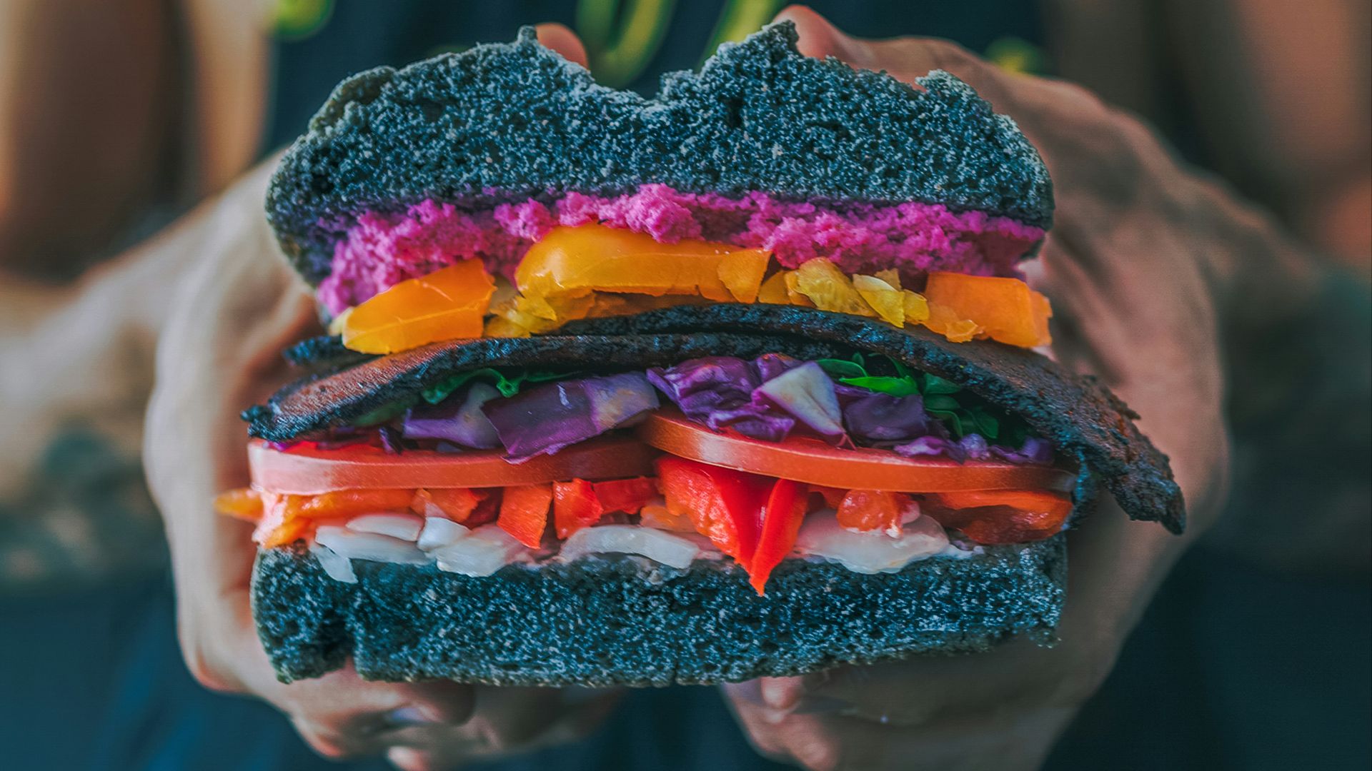 person holding meat sandwich with sliced tomatoes, onions, and grilled meat