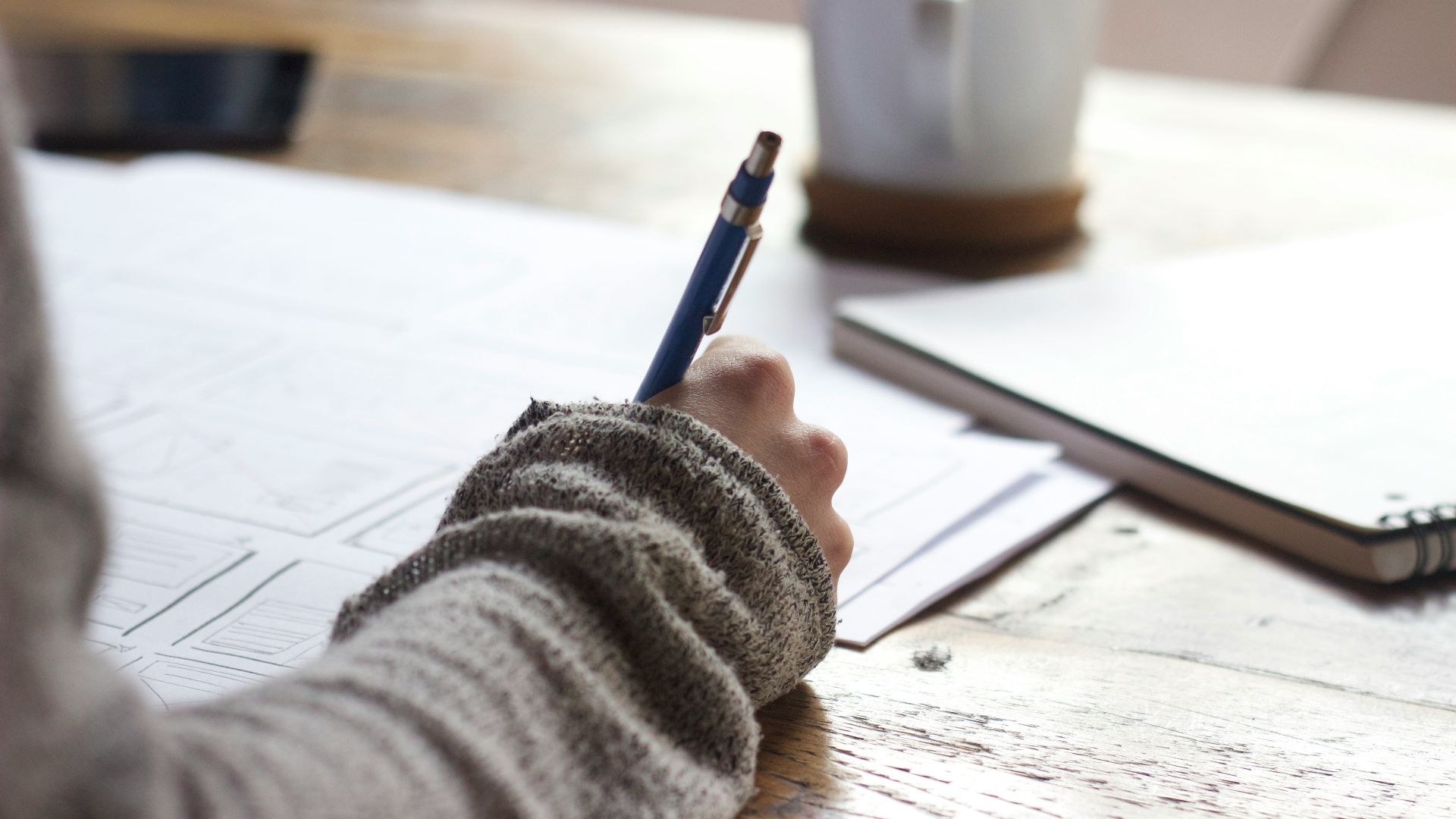 person writing on brown wooden table near white ceramic mug