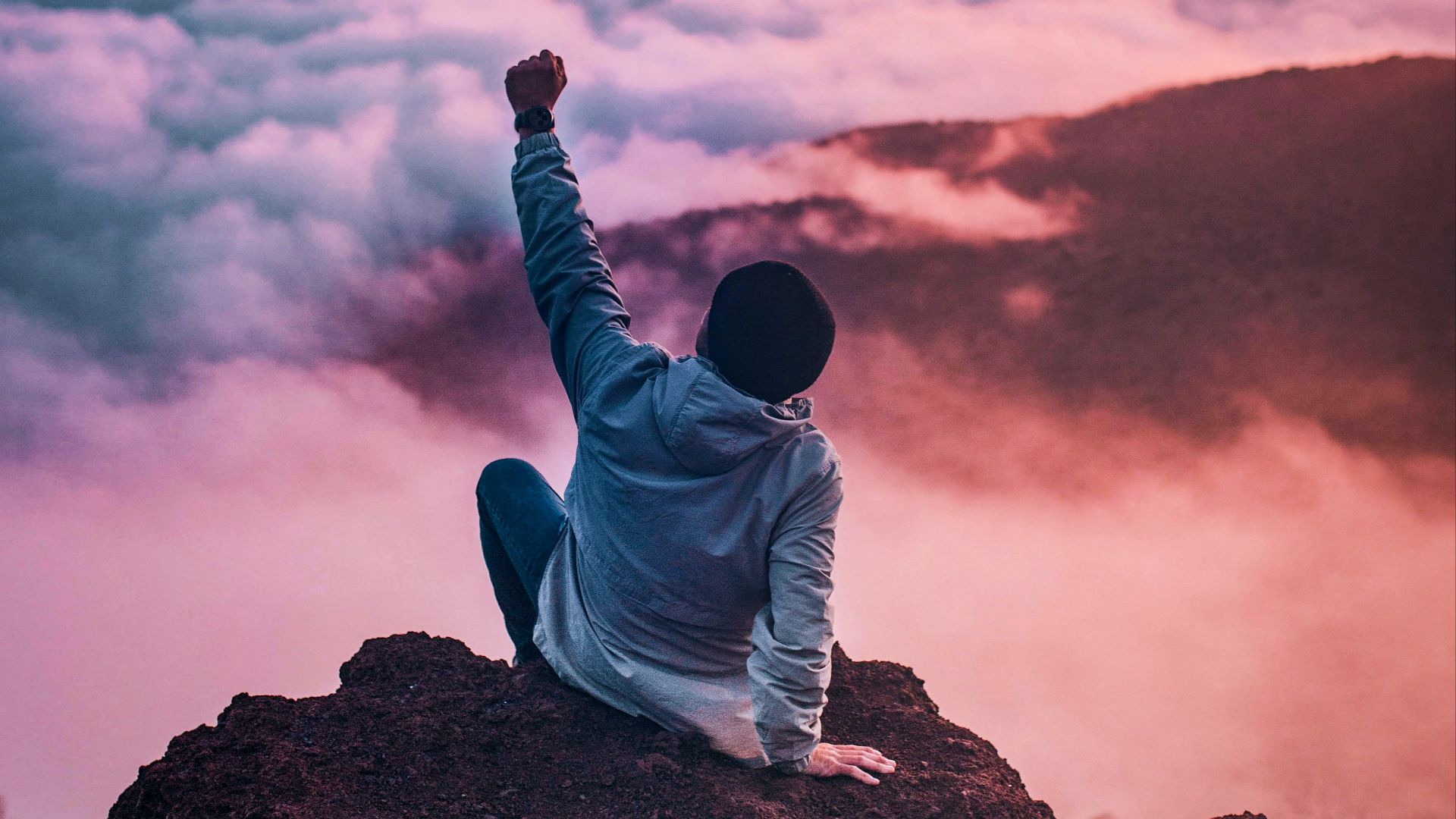 man sitting on mountain cliff facing white clouds rising one hand at golden hour
