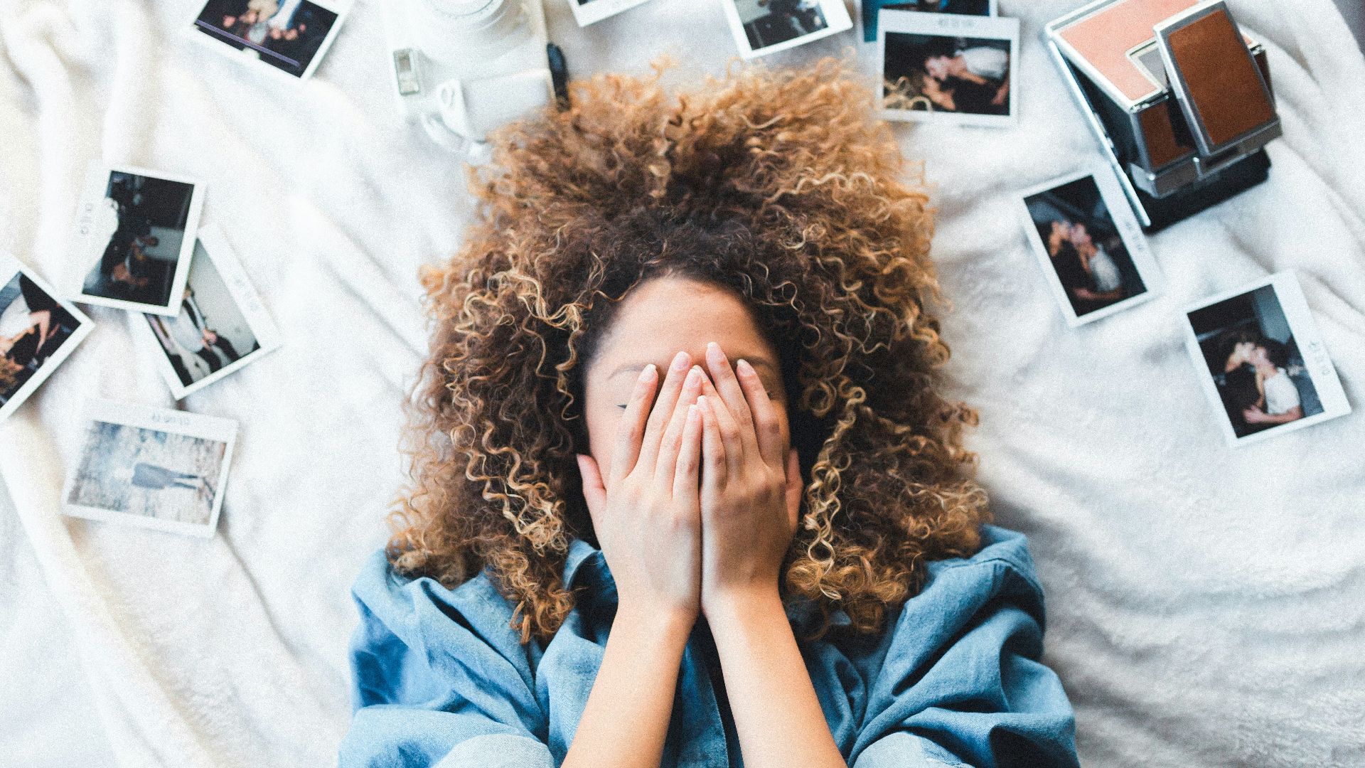 woman lying on bed covering her face surrounded by photos and white camera