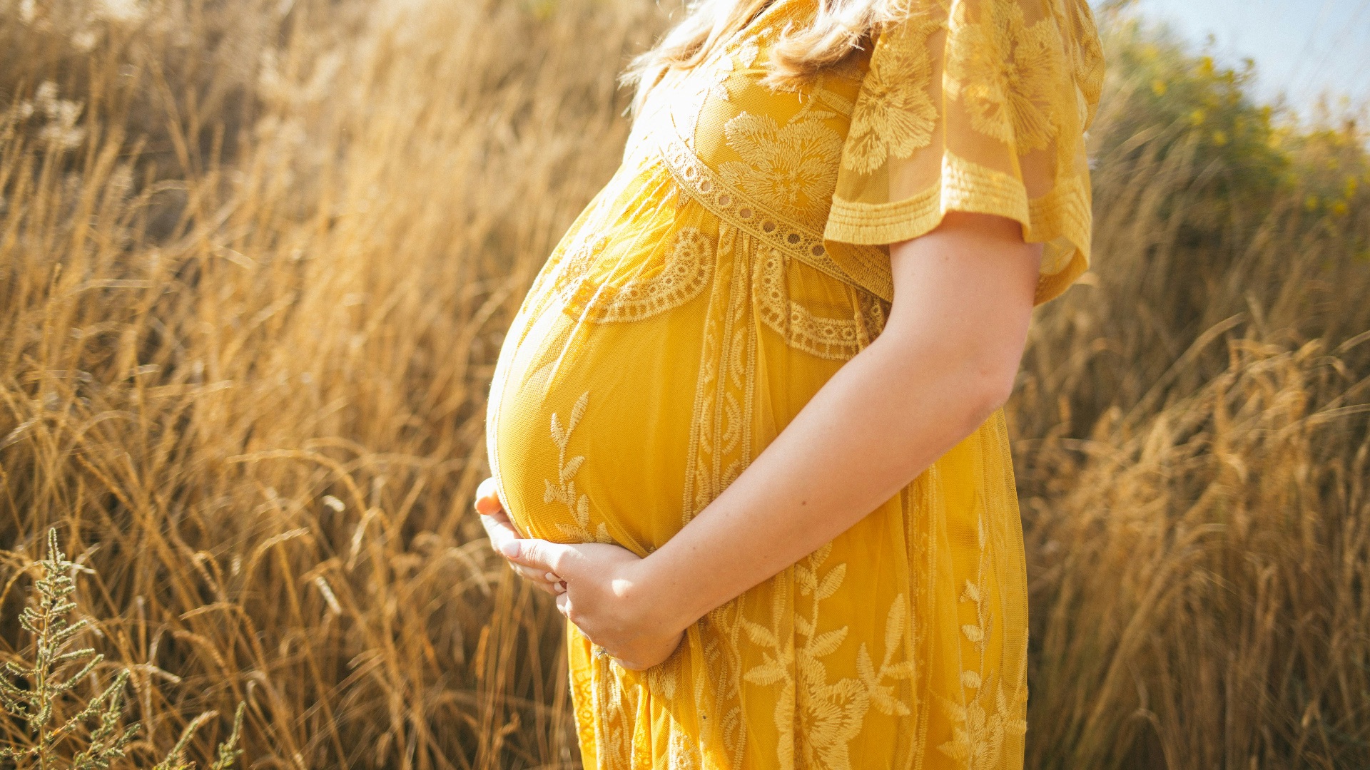 pregnant woman wearing yellow floral dress standing while touching her tummy and facing her right side near brown field during daytime