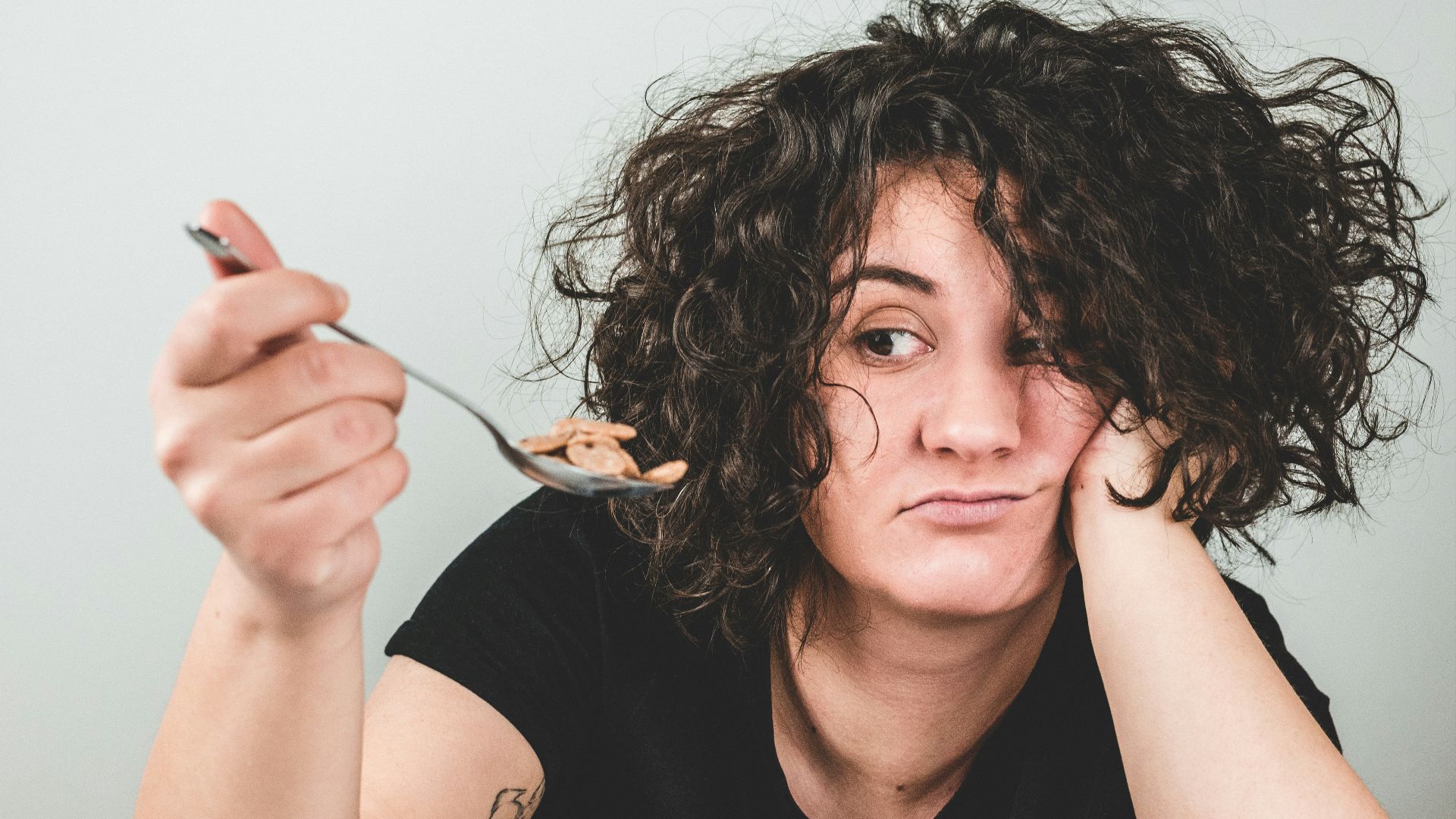 woman with messy hair wearing black crew-neck t-shirt holding spoon with cereals on top