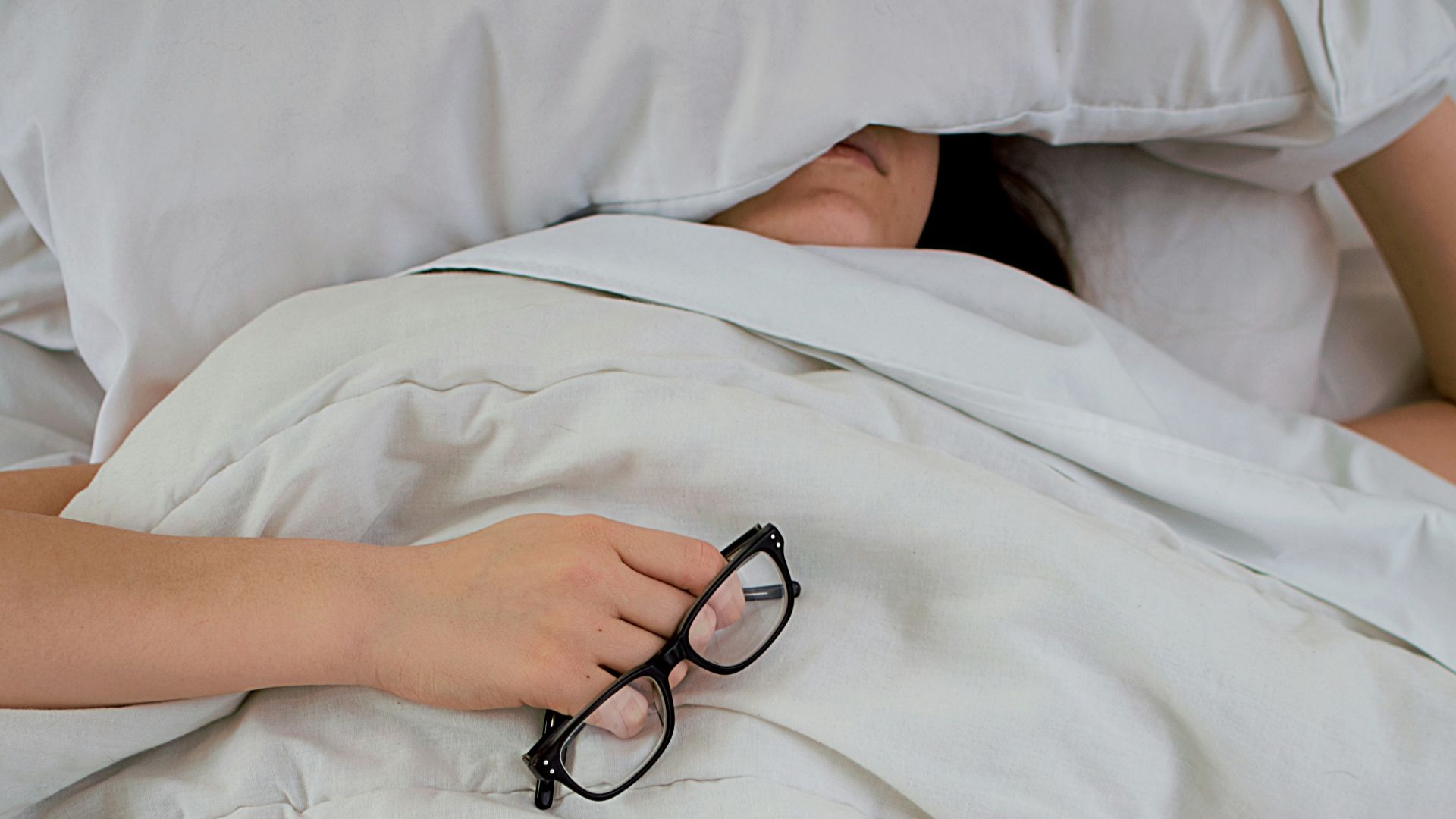 person lying on bed while covering face with pillow and holding eyeglasses