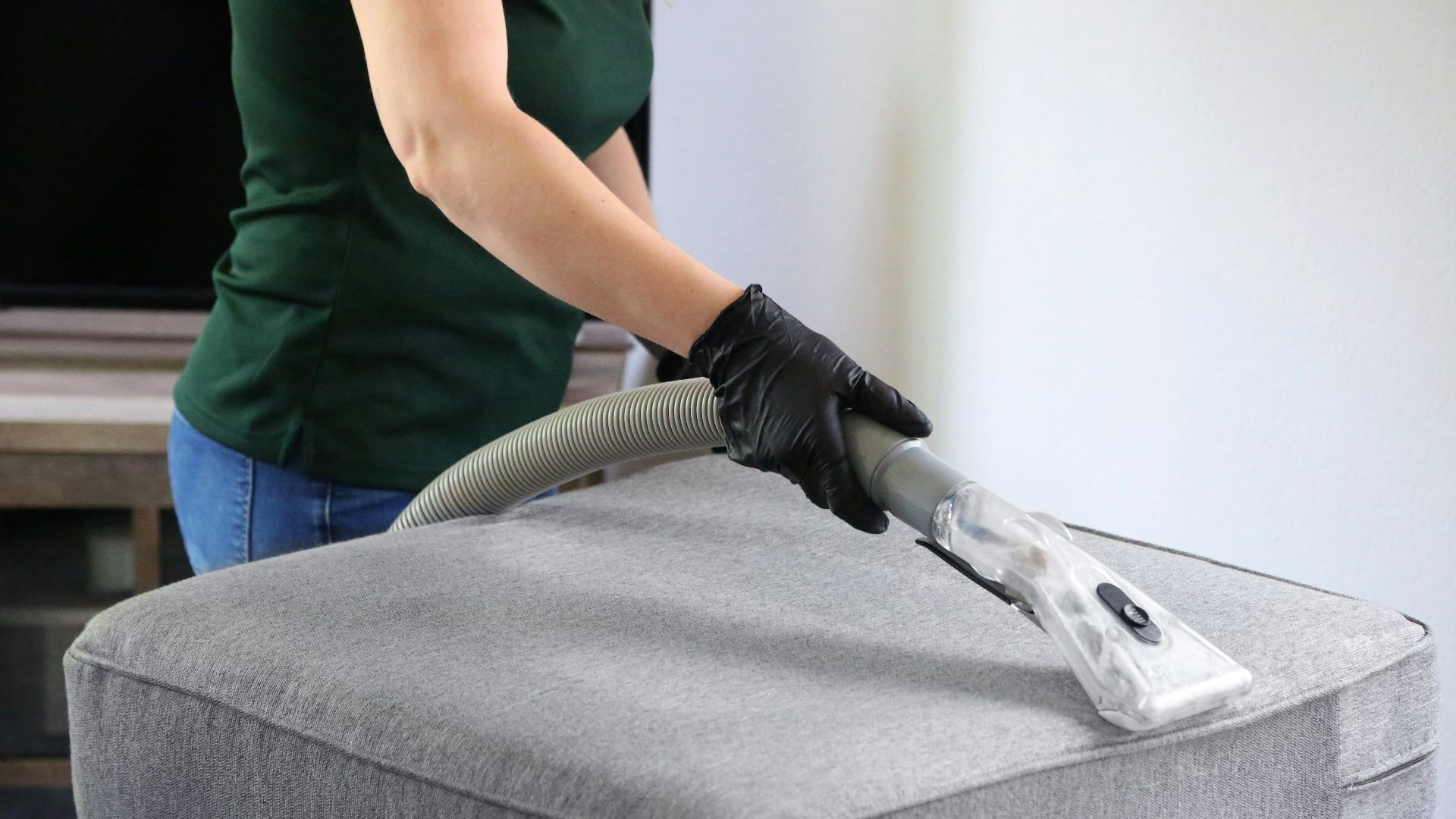 a woman in a green shirt and black gloves vacuuming a gray ottoman