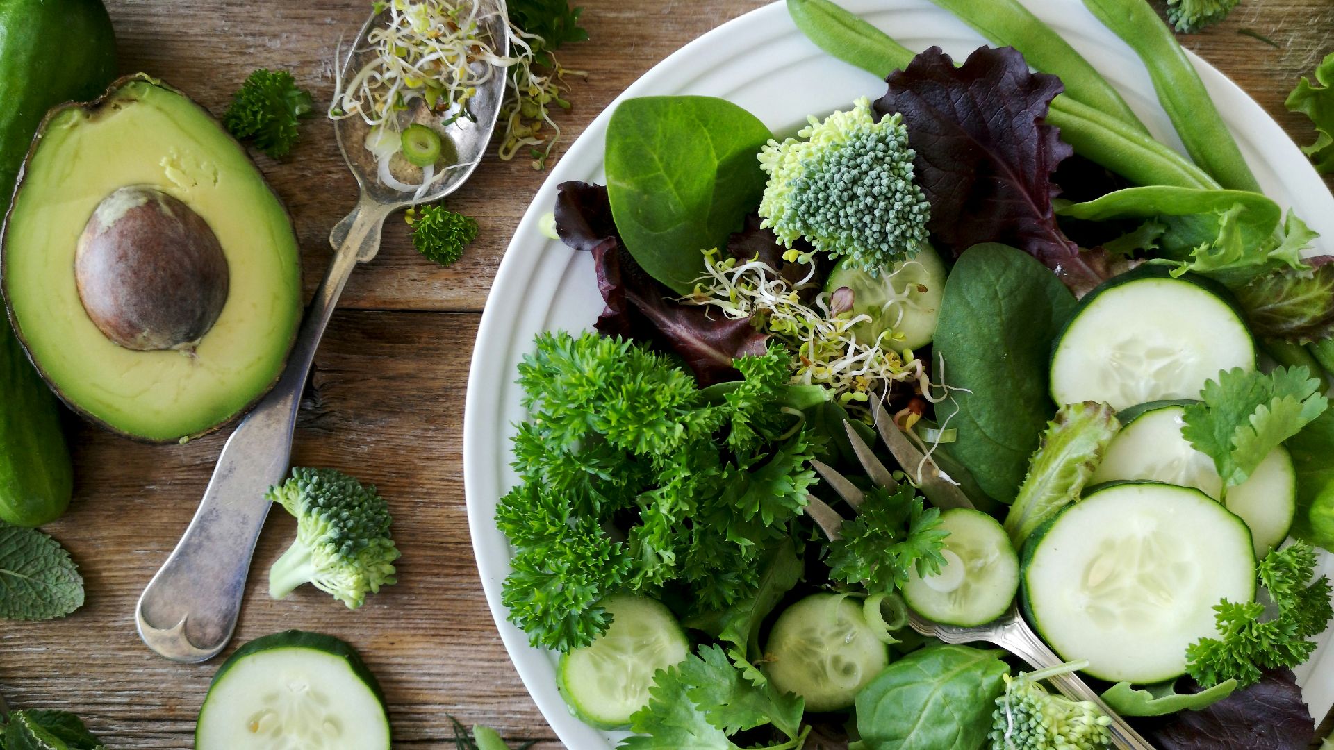sliced broccoli and cucumber on plate with gray stainless steel fork near green bell pepper, snowpea, and avocado fruit