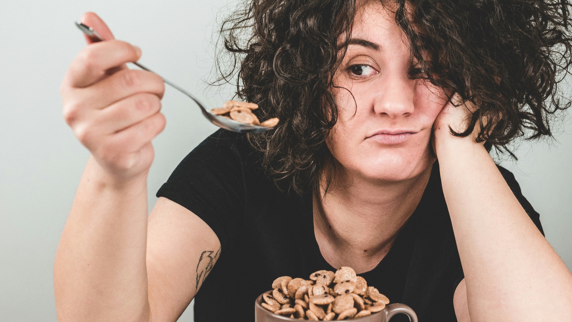 woman with messy hair wearing black crew-neck t-shirt holding spoon with cereals on top