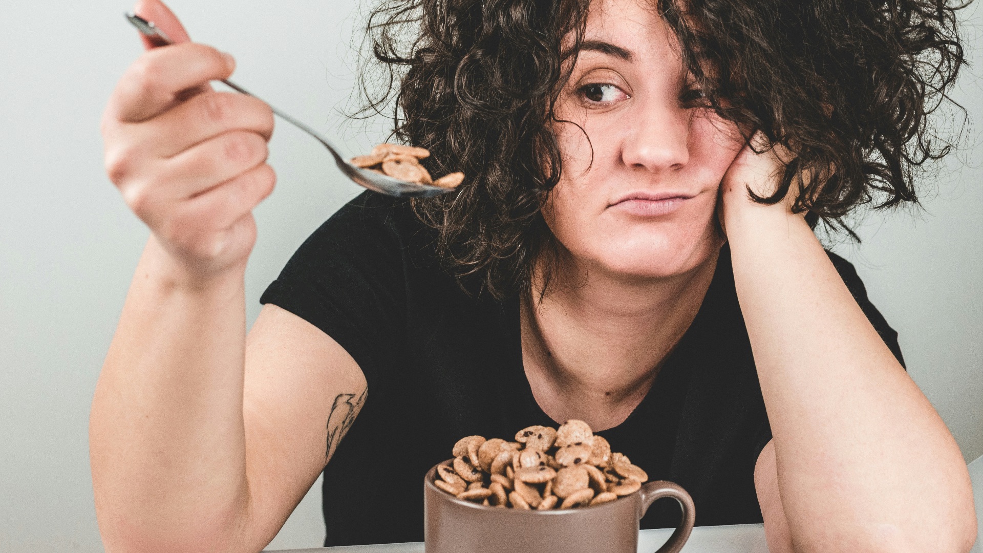 woman with messy hair wearing black crew-neck t-shirt holding spoon with cereals on top