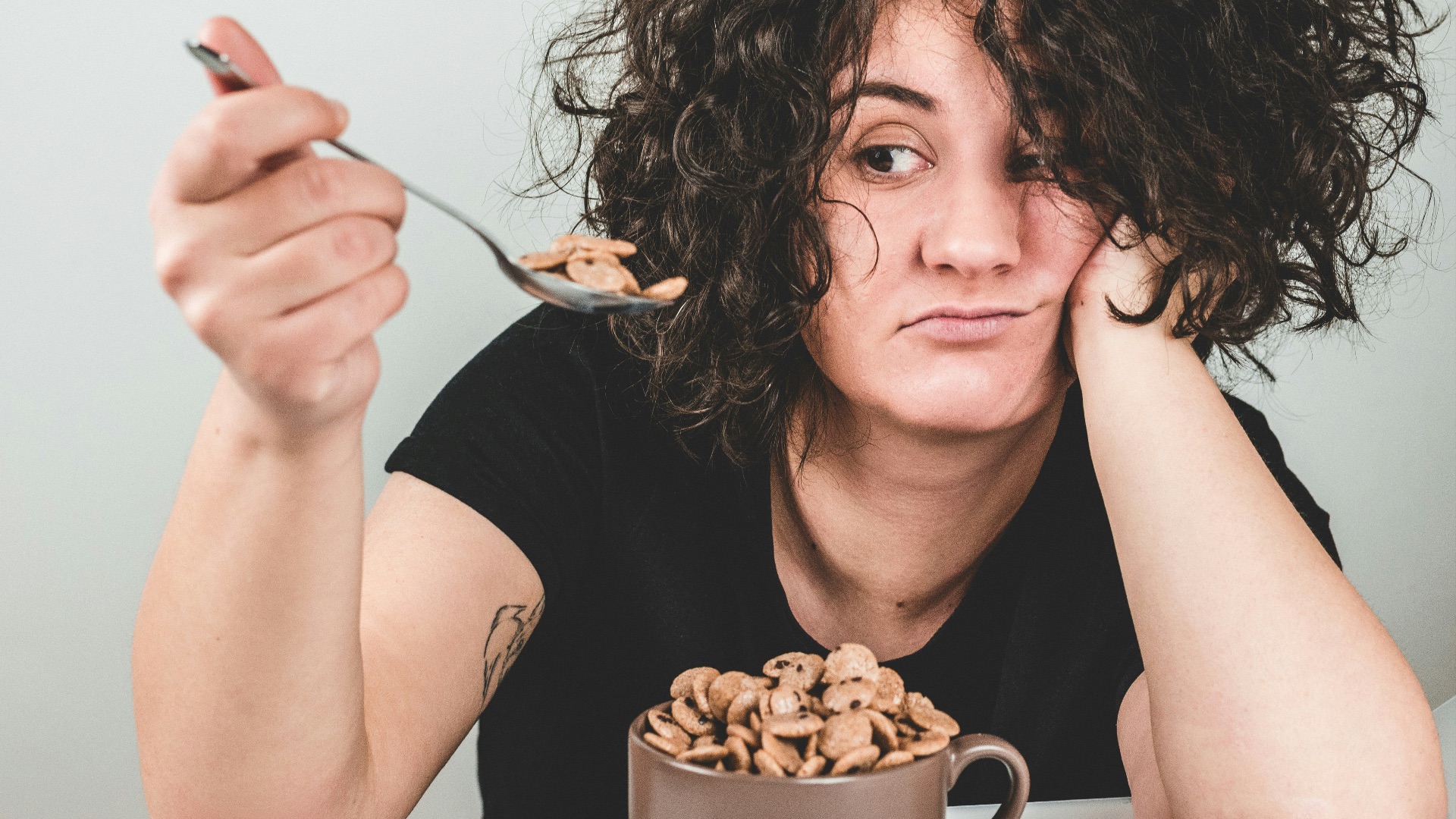 woman with messy hair wearing black crew-neck t-shirt holding spoon with cereals on top