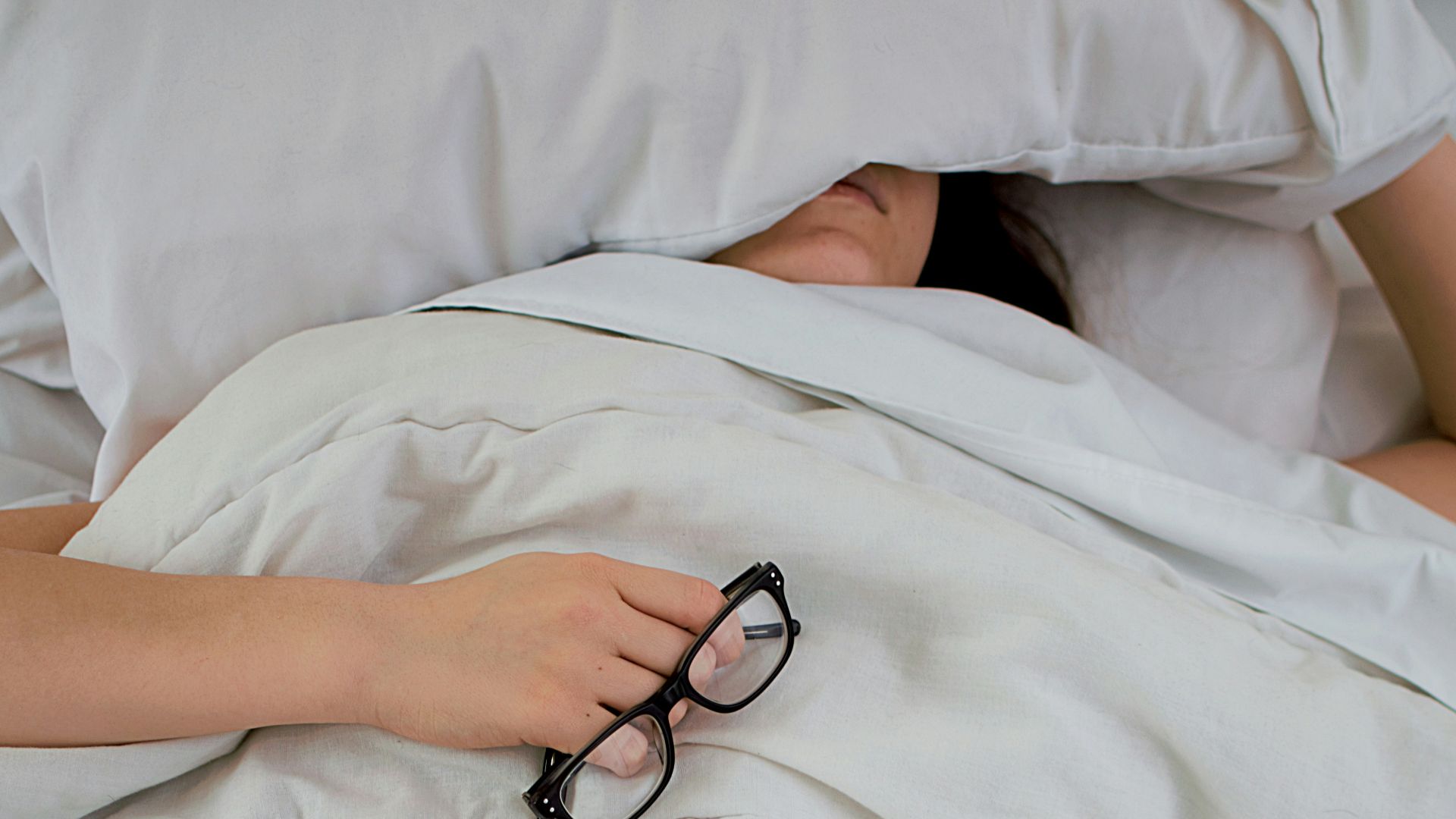 person lying on bed while covering face with pillow and holding eyeglasses
