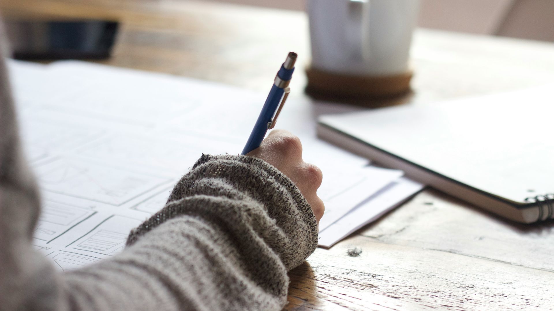 person writing on brown wooden table near white ceramic mug