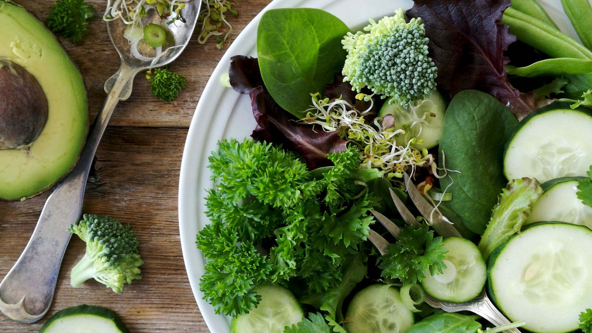 sliced broccoli and cucumber on plate with gray stainless steel fork near green bell pepper, snowpea, and avocado fruit
