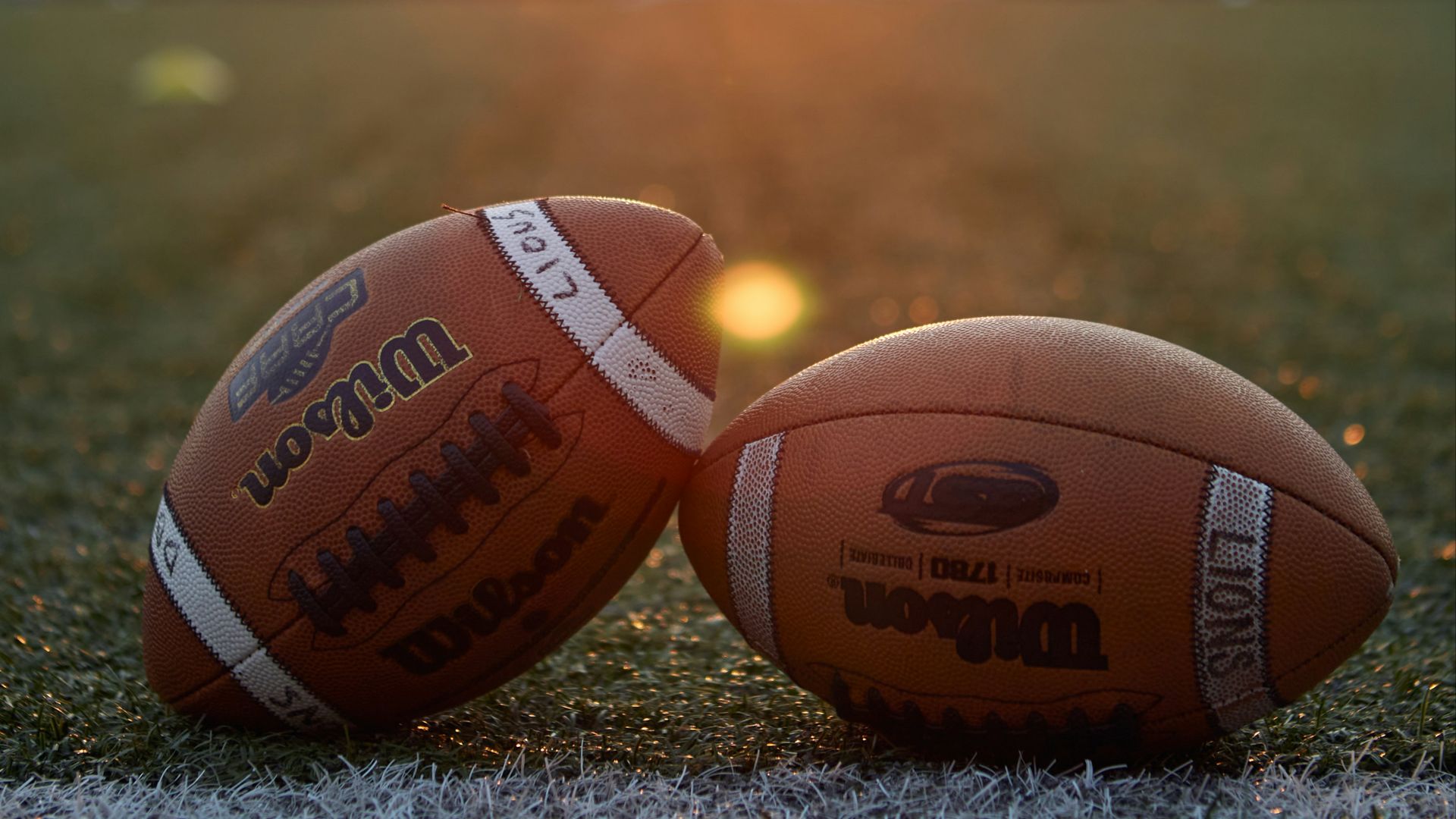 Two footballs sitting on a football field at sunset