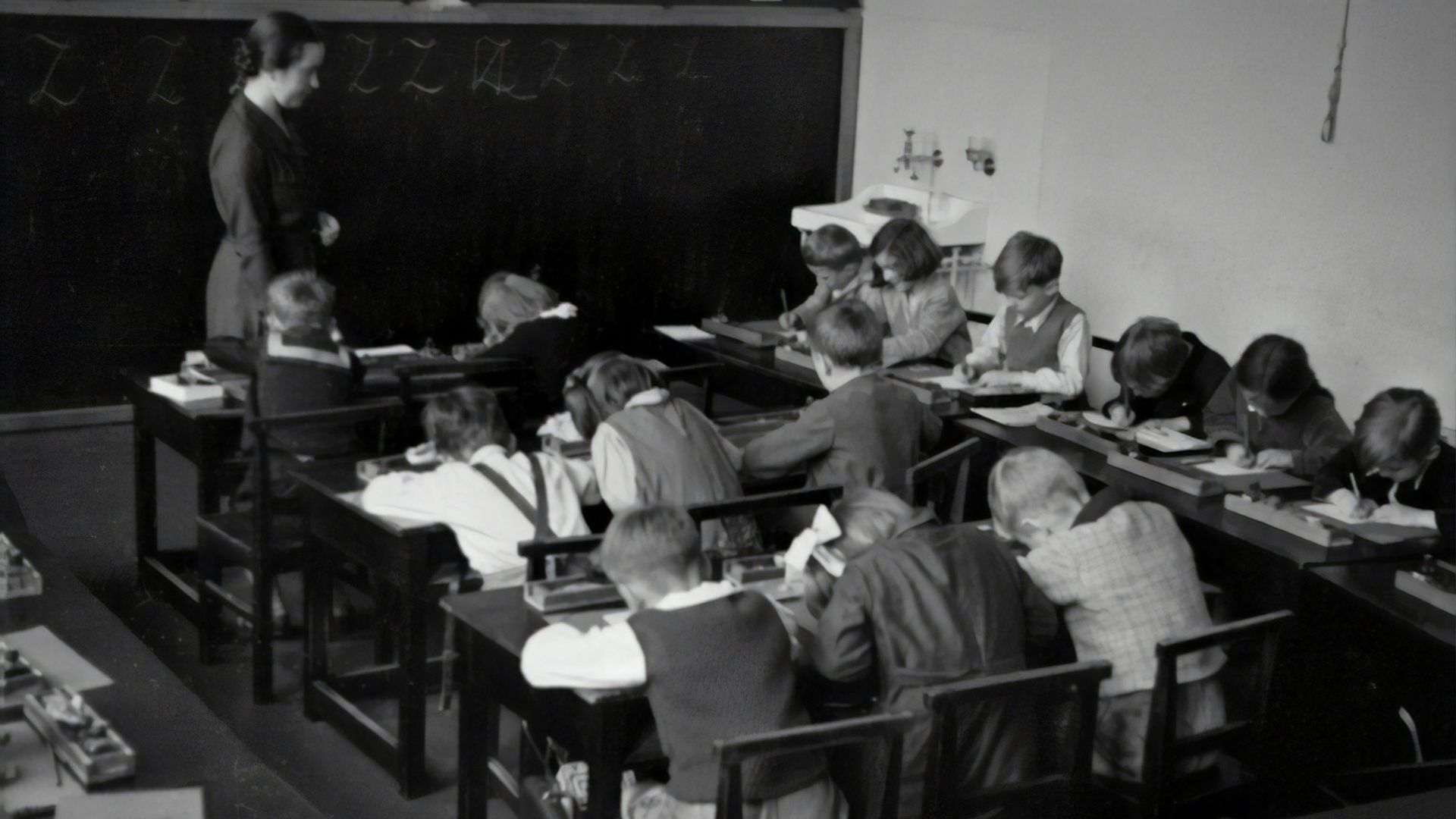 grayscale photography of teacher standing near chalkboard and children sitting on chairs