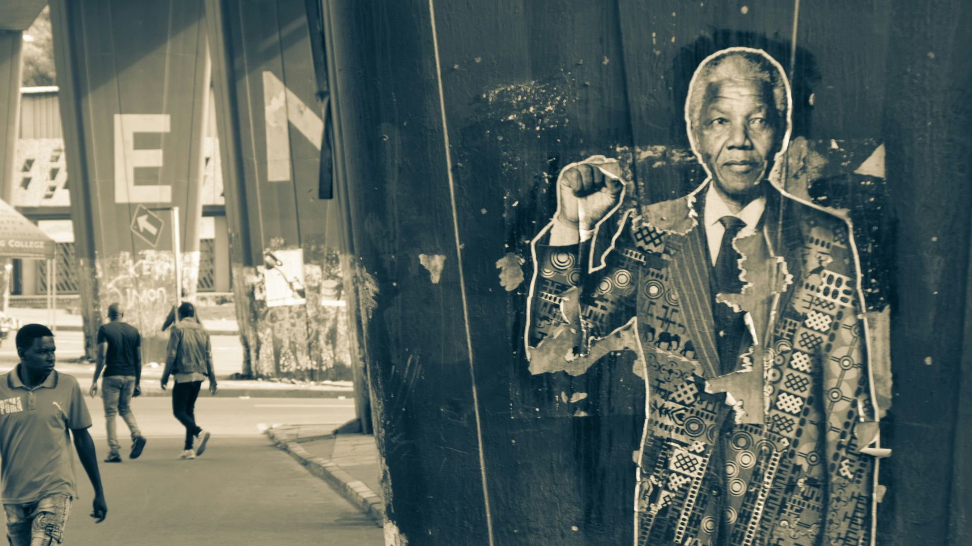 grayscale photo of man in black and white camouflage jacket standing beside wall
