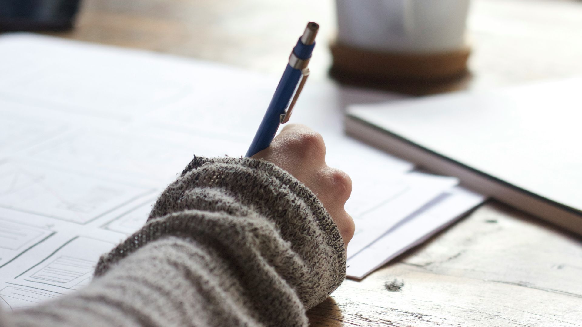 person writing on brown wooden table near white ceramic mug