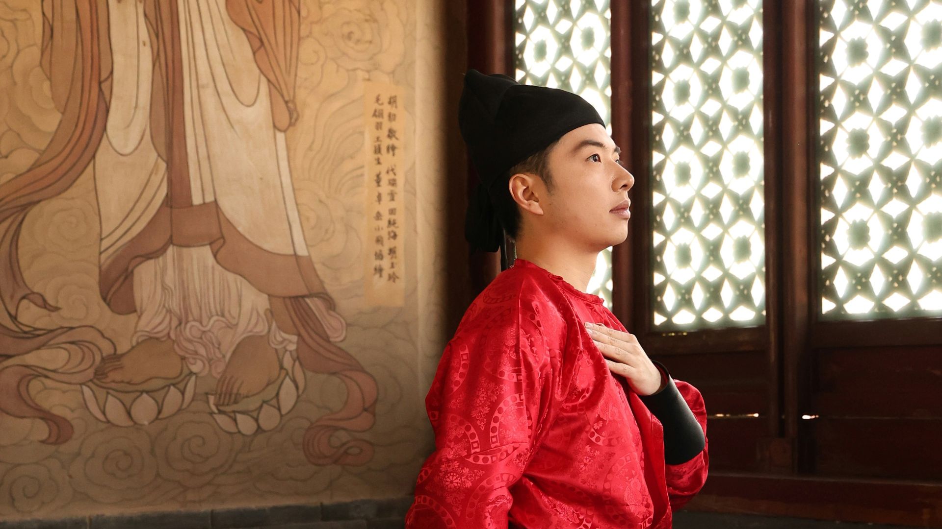 Man in red traditional chinese attire stands by mural.
