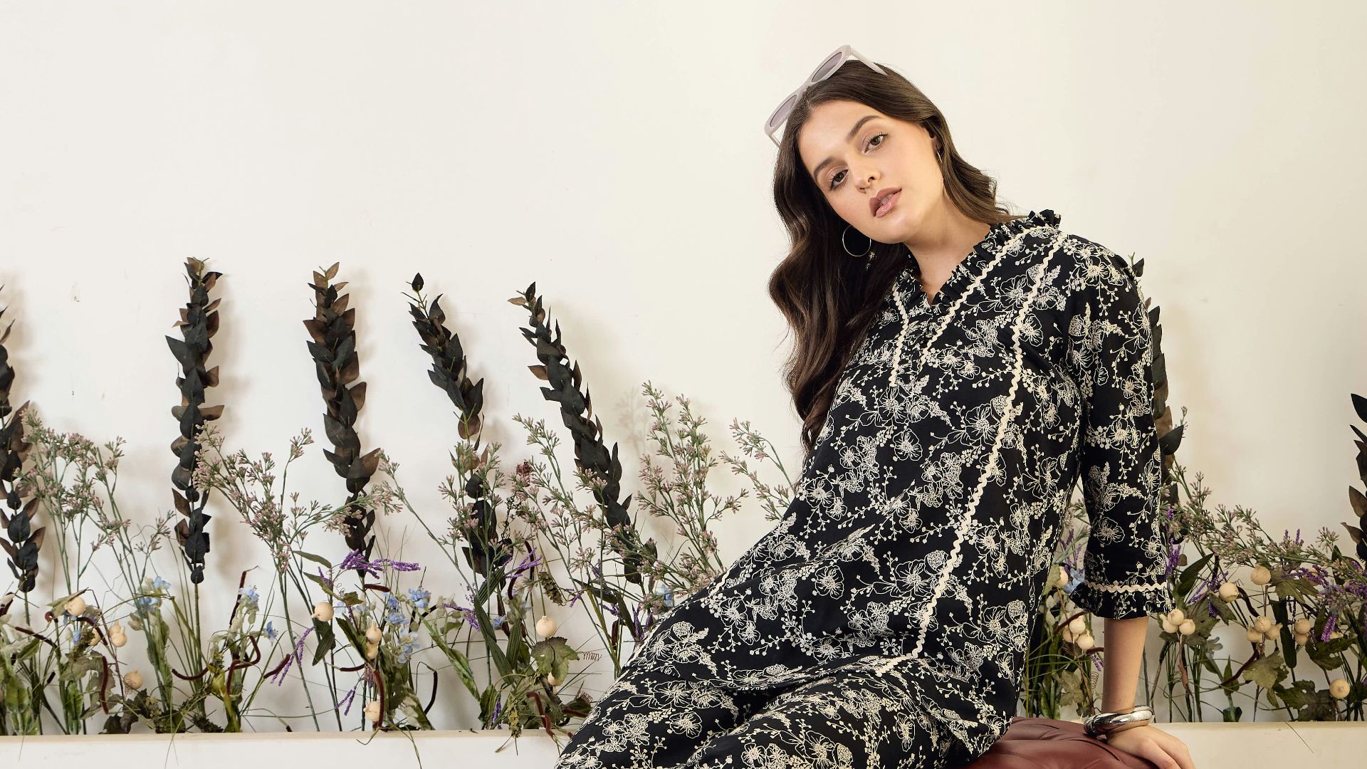 Woman in floral jumpsuit sitting on stool.