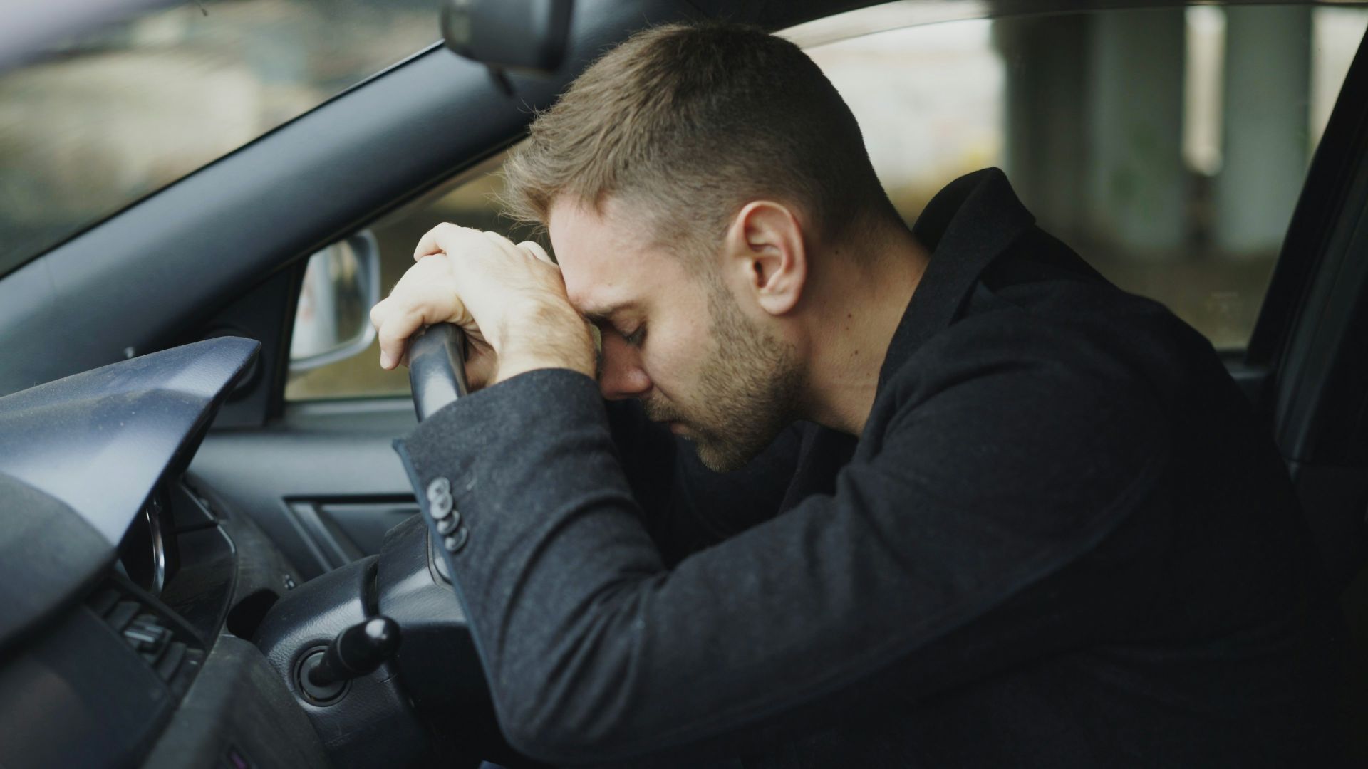 Man resting head on steering wheel in car.