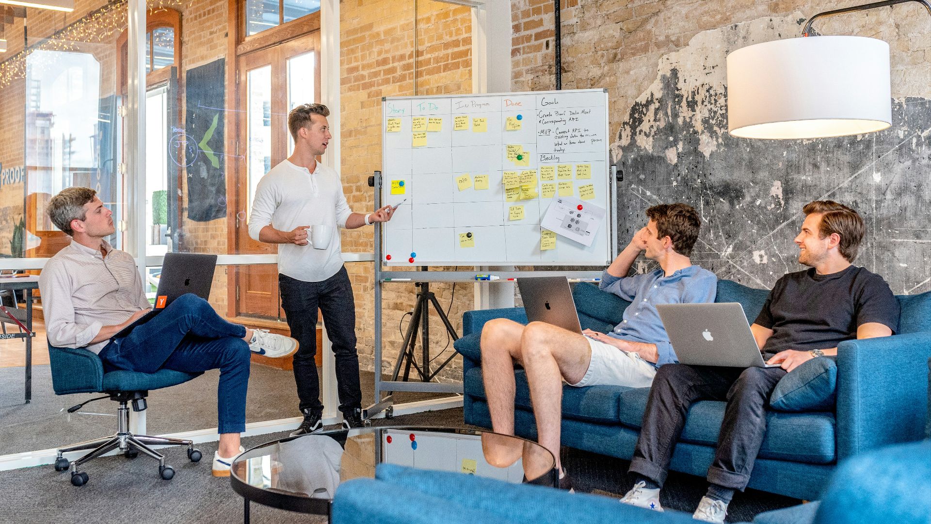 three men sitting while using laptops and watching man beside whiteboard