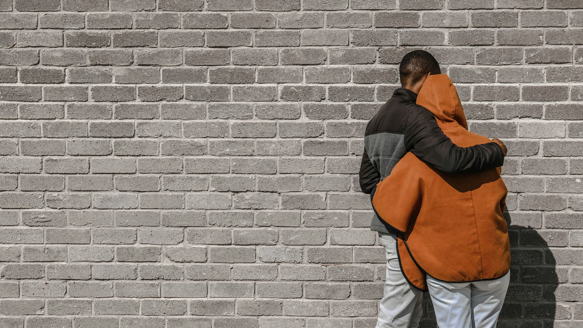 man in black and brown jacket and white pants standing beside brick wall