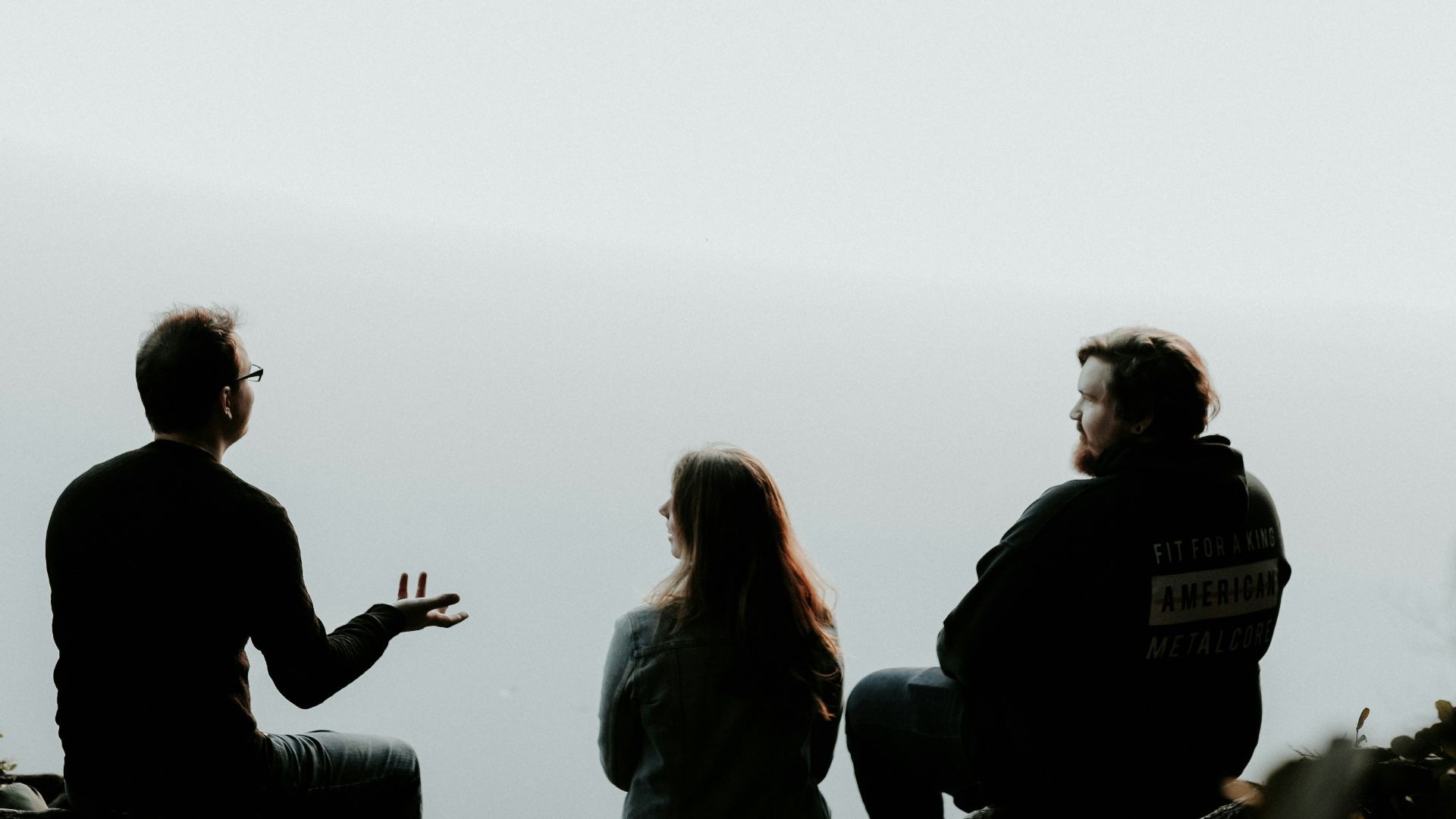 silhouette of three people sitting on cliff under foggy weather
