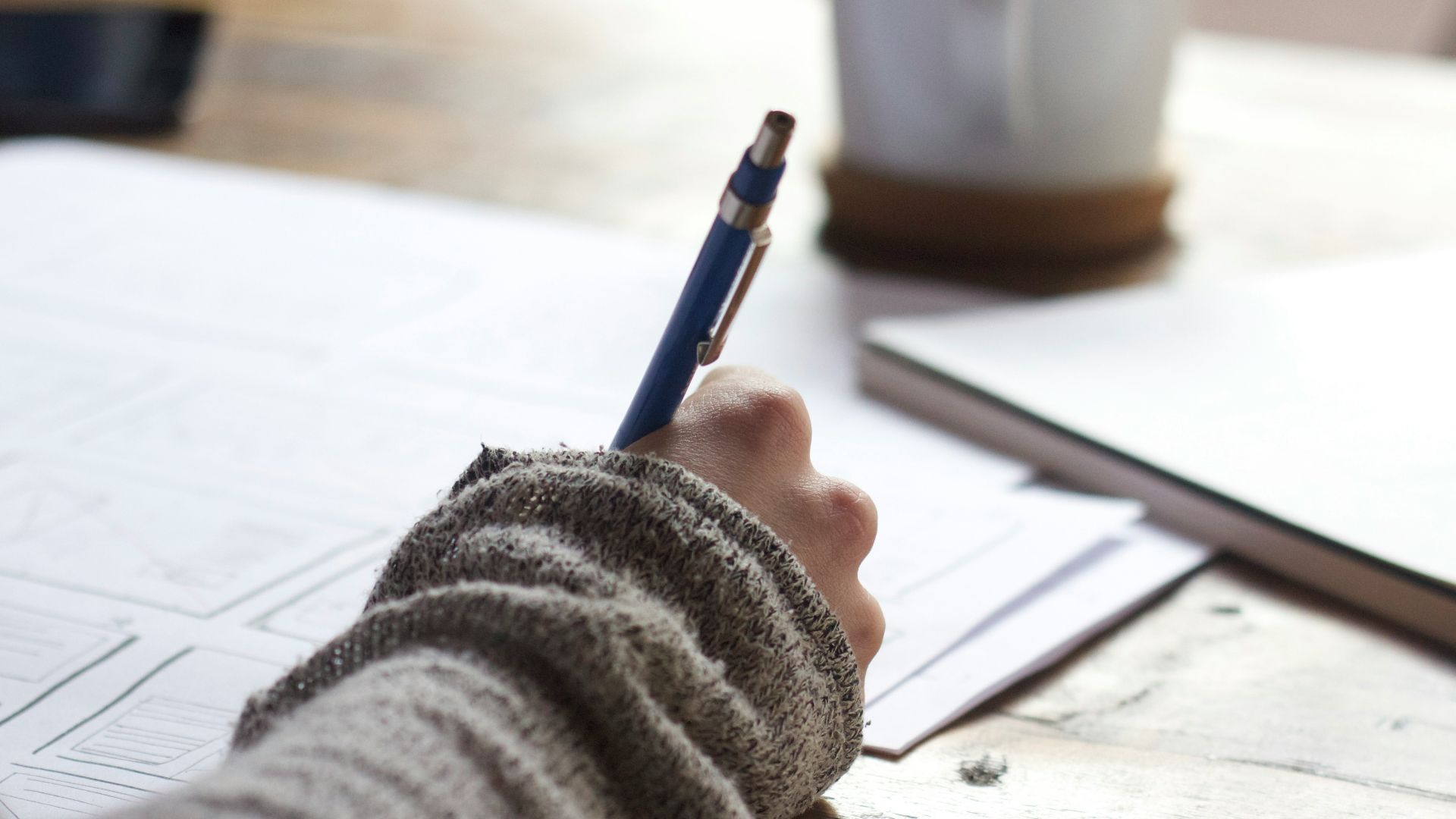 person writing on brown wooden table near white ceramic mug