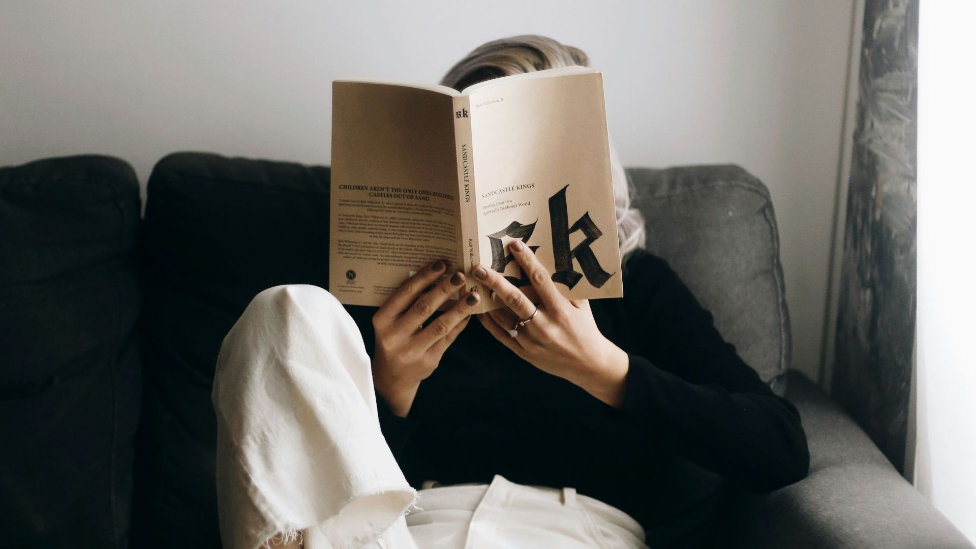 woman in white shirt reading book sitting on black sofa