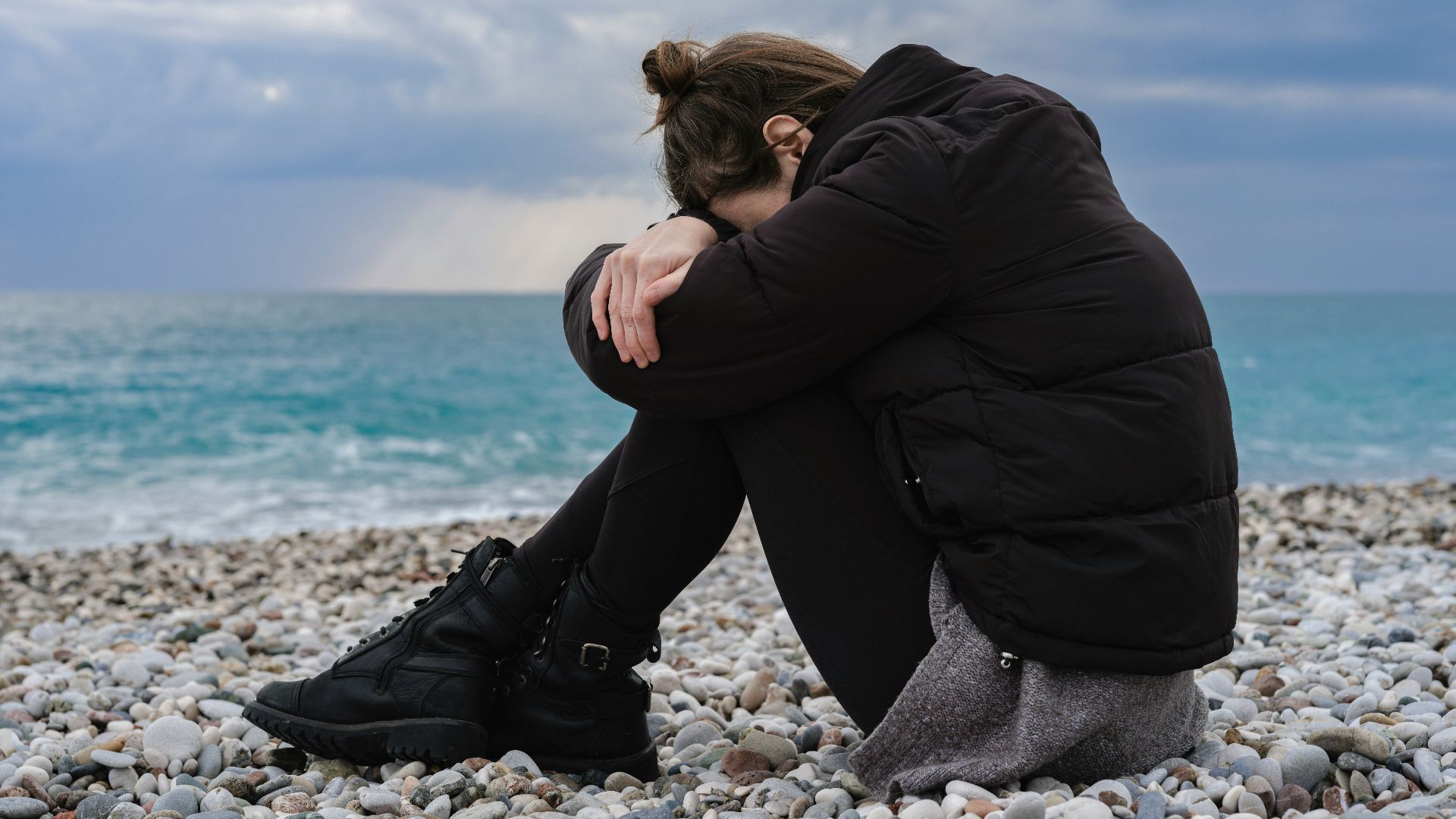 a woman sitting on a rocky beach with her head in her hands