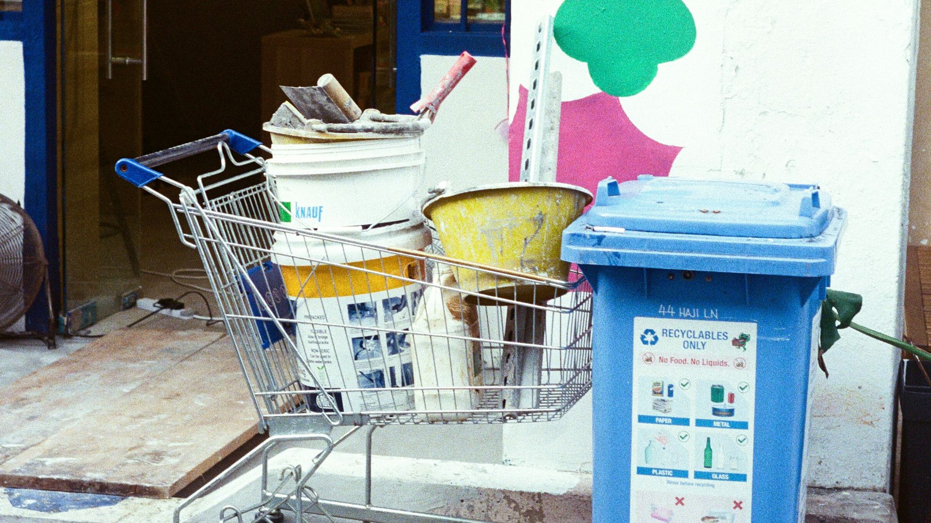 Shopping cart filled with painting supplies next to trash bin.