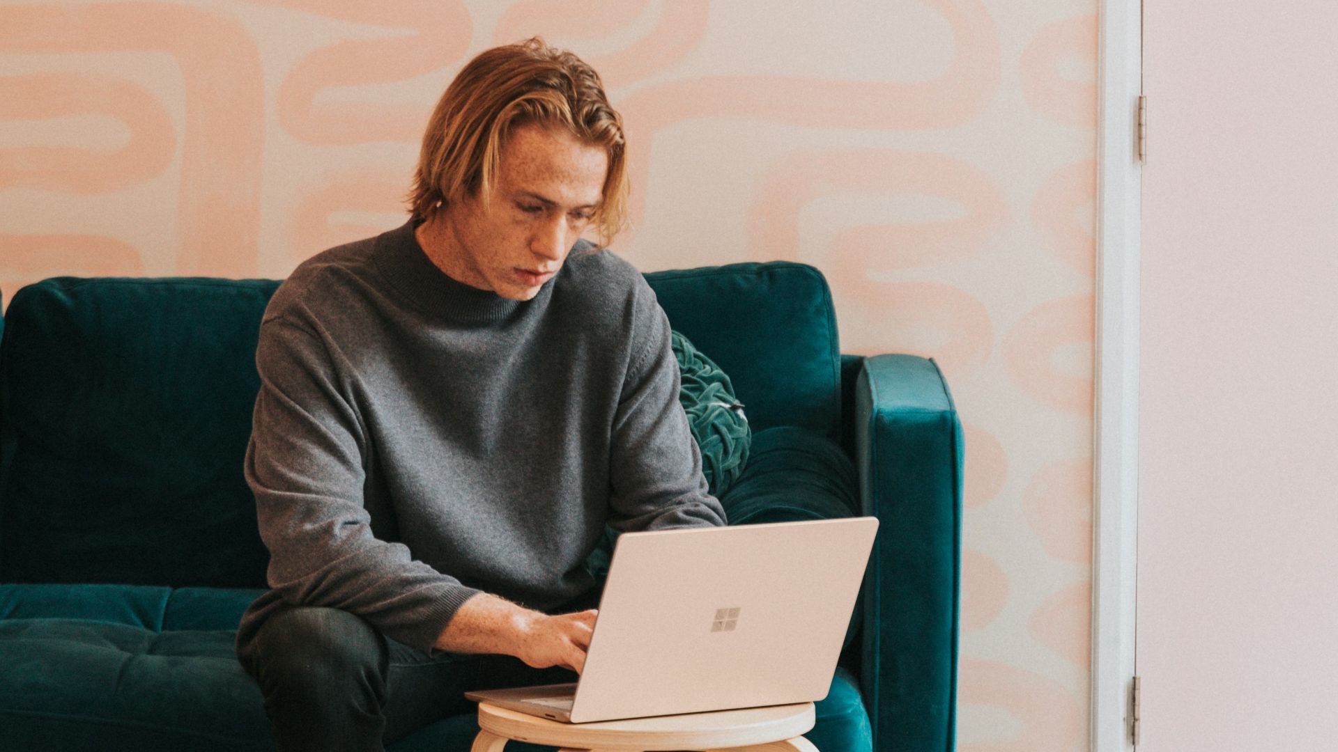 man in gray long sleeve shirt sitting on blue sofa working on a Microsoft Surface laptop