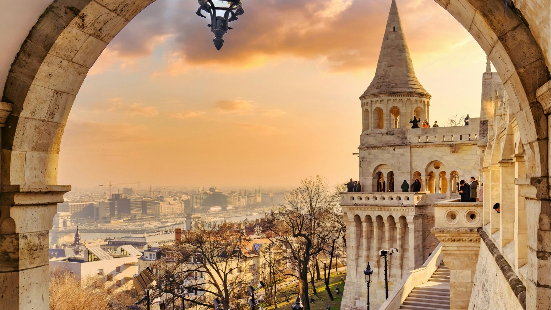 a stone walkway with a lamp post and a large stone arch with a city in the background