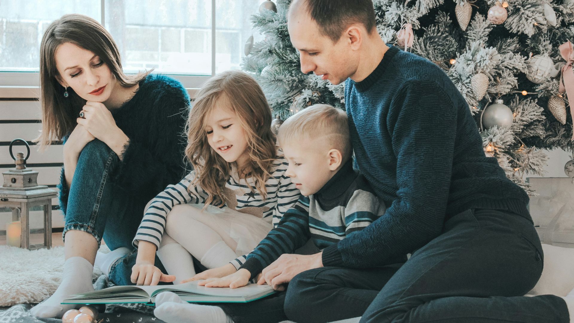 girl and boy reading book sitting between man and woman beside Christmas tree
