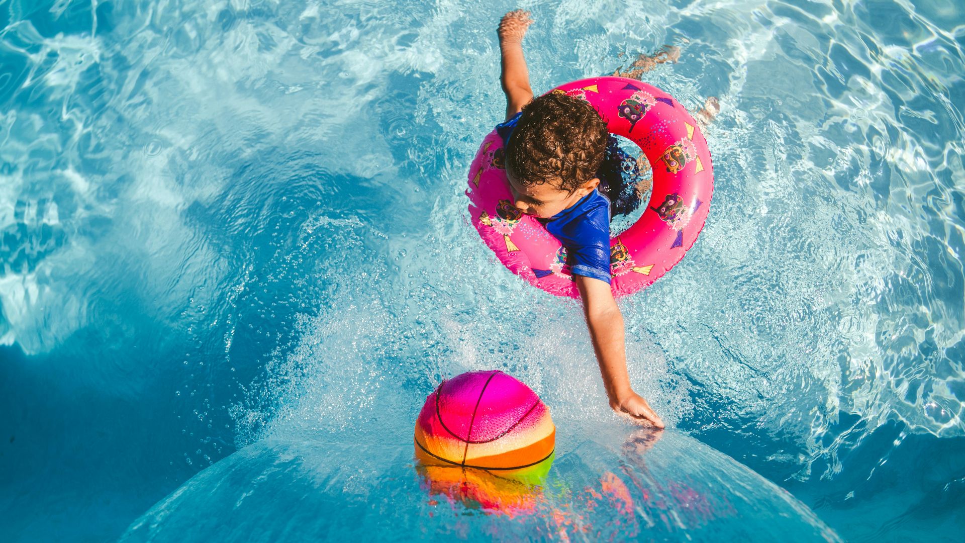 girl in blue tank top and blue shorts holding pink inflatable ring on water