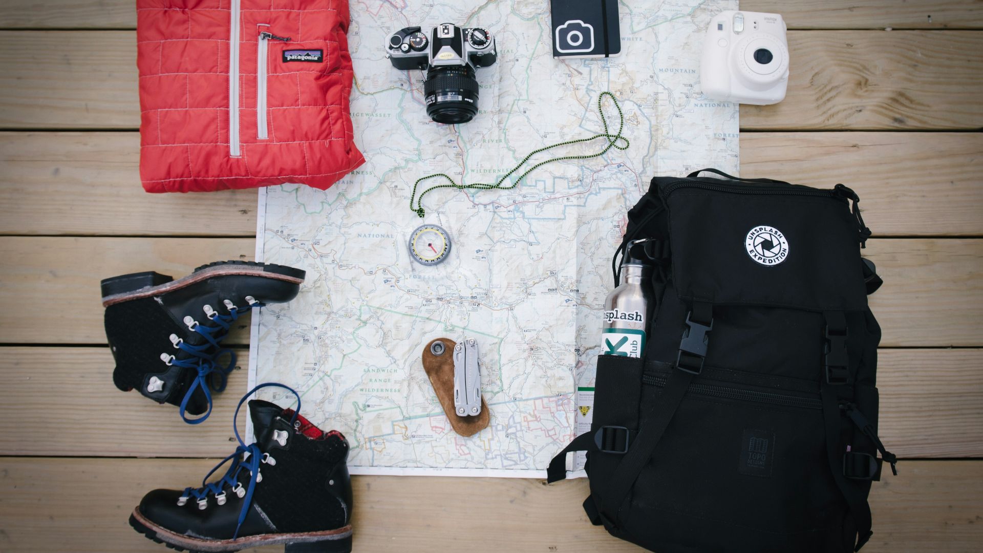 black hiking backpack near white Fujifilm instax mini camera near black leather boots, red half-zip jacket, gray pocket watch on white map