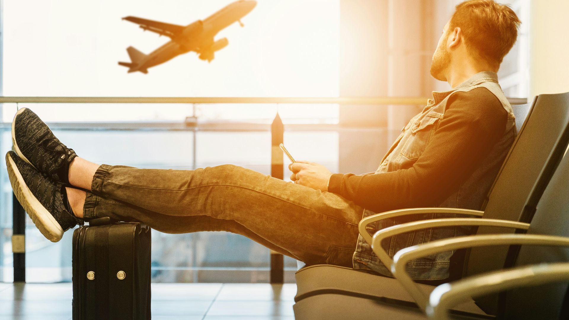 man sitting on gang chair with feet on luggage looking at airplane