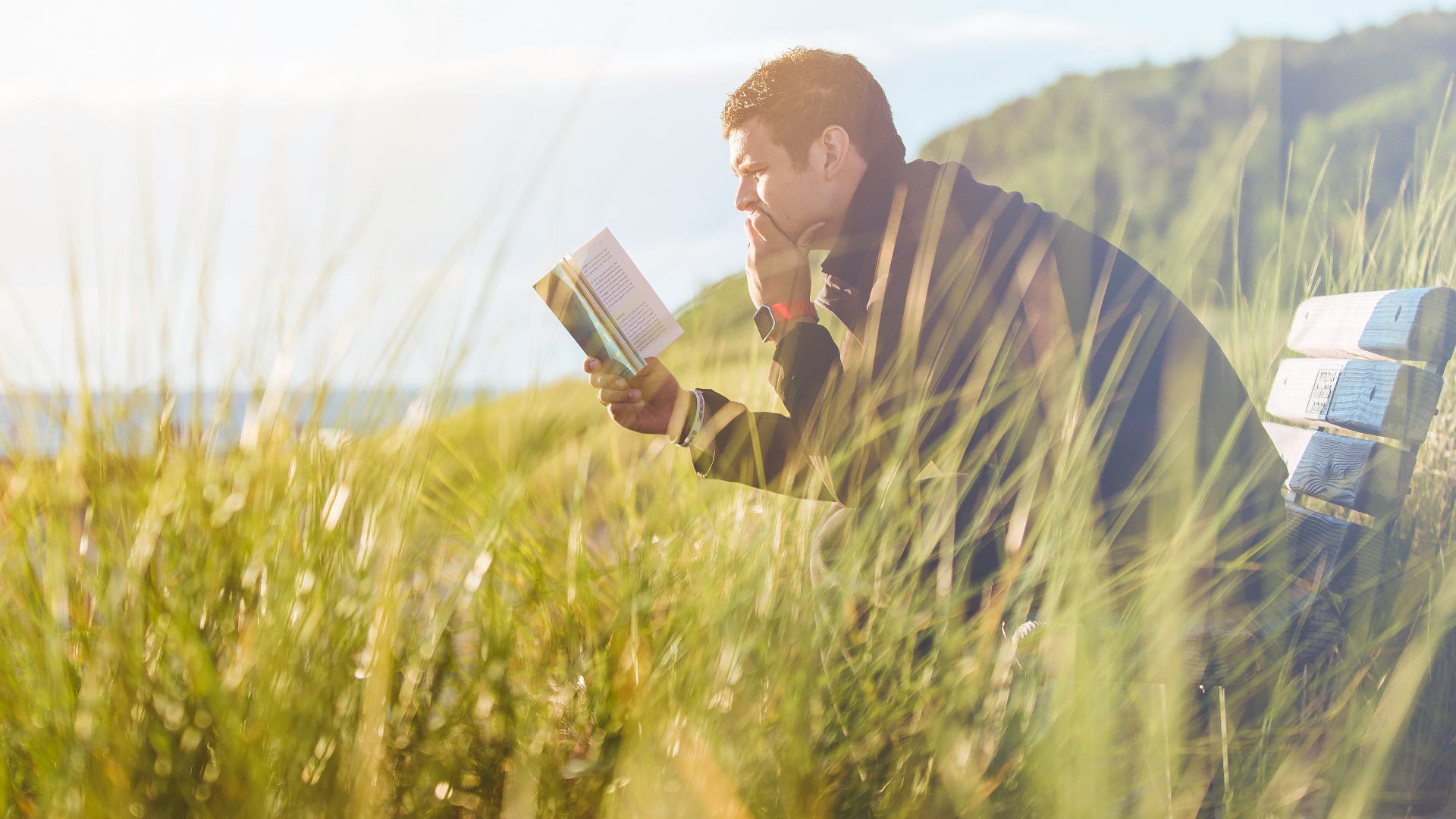man reading book on beach near lake during daytime