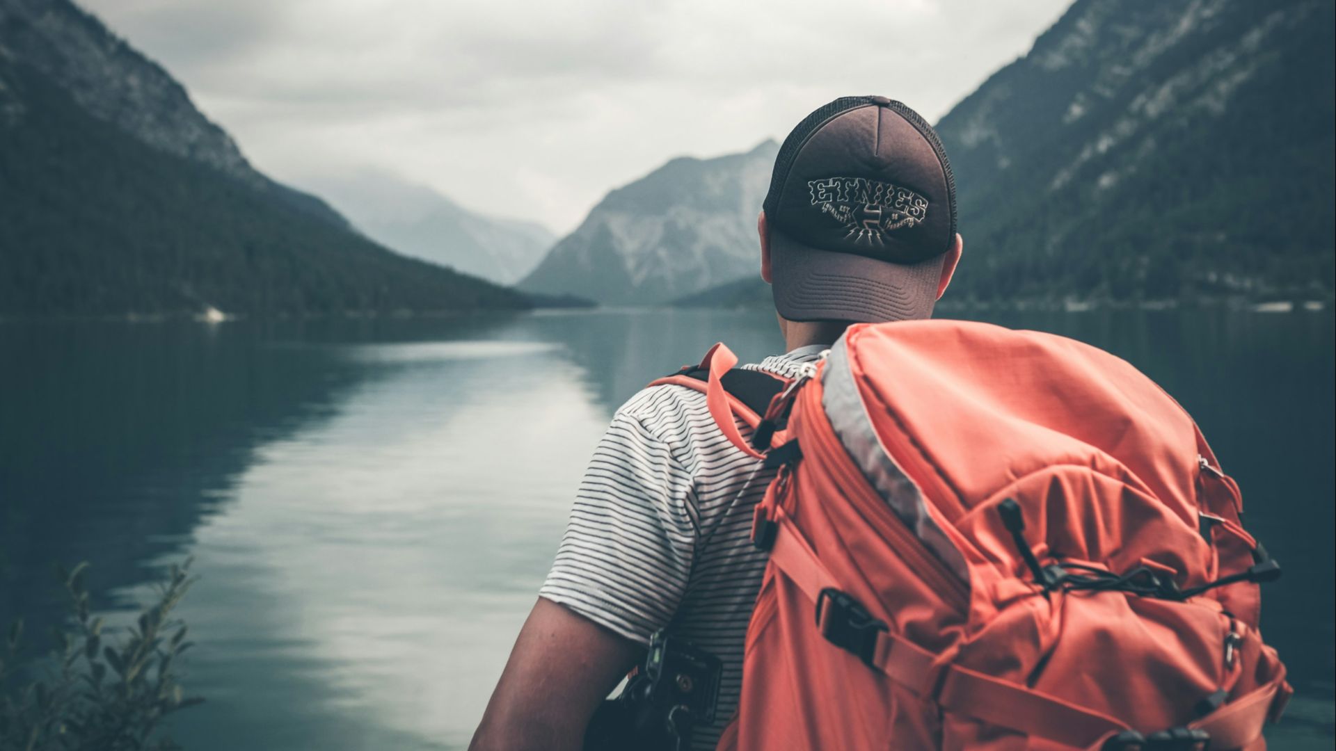 man with red hiking backpack facing body of water and mountains at daytime
