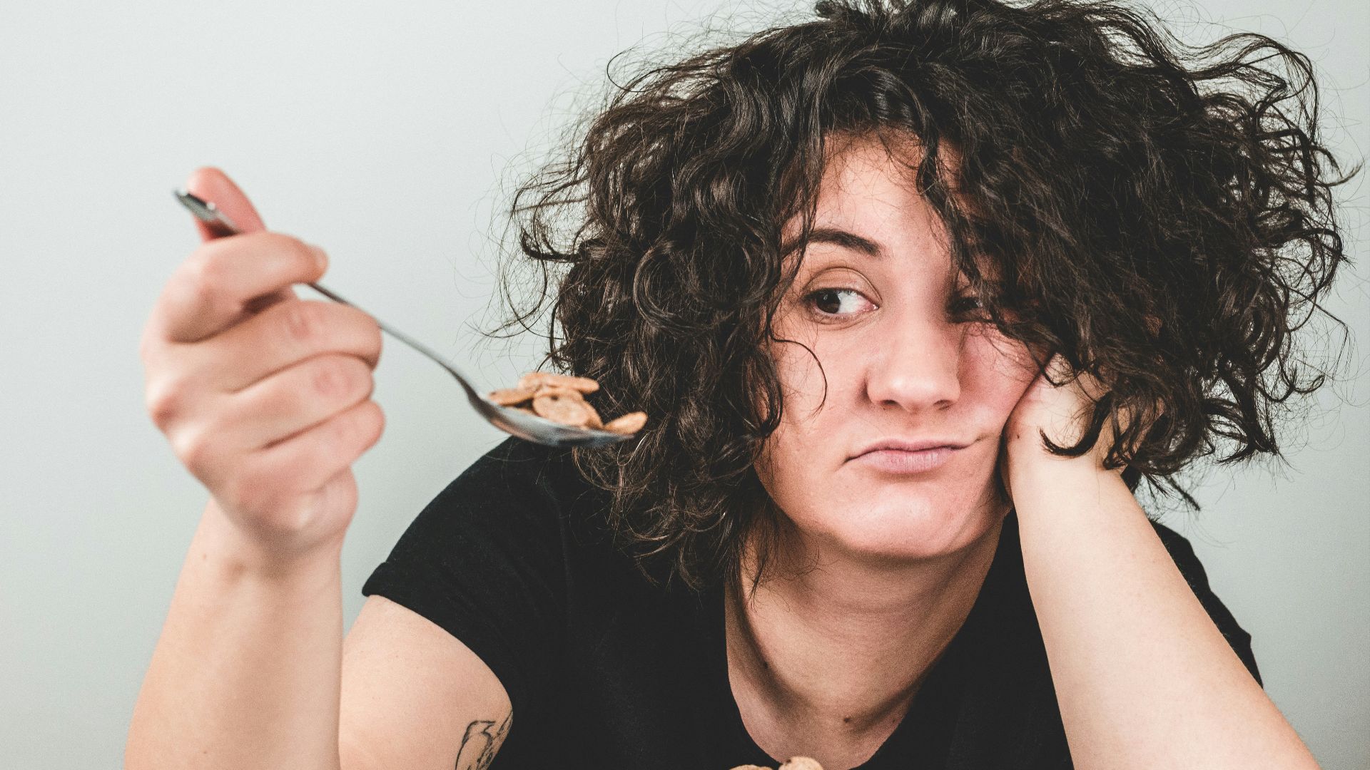 woman with messy hair wearing black crew-neck t-shirt holding spoon with cereals on top