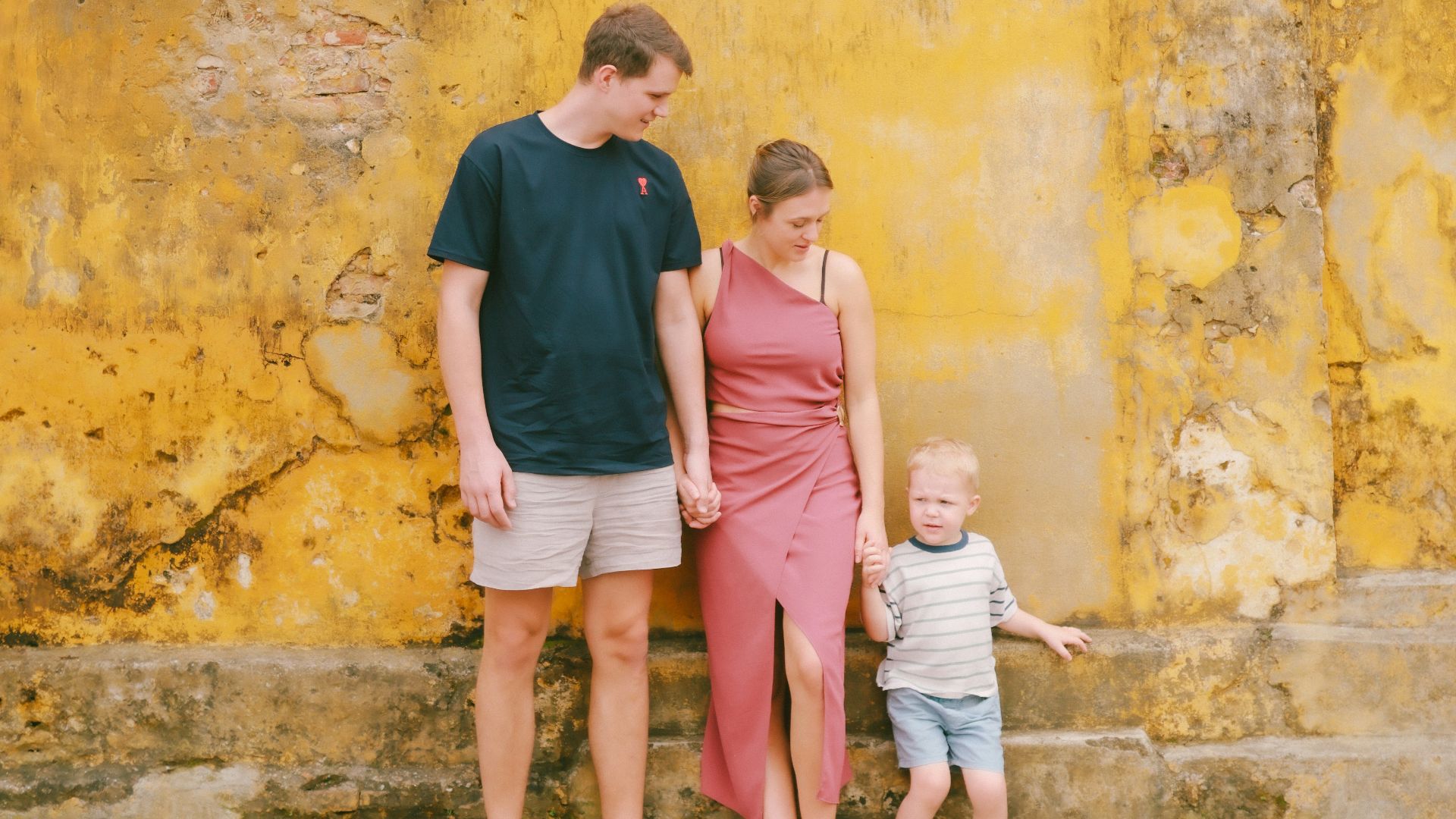 Family of three standing against a textured yellow wall.