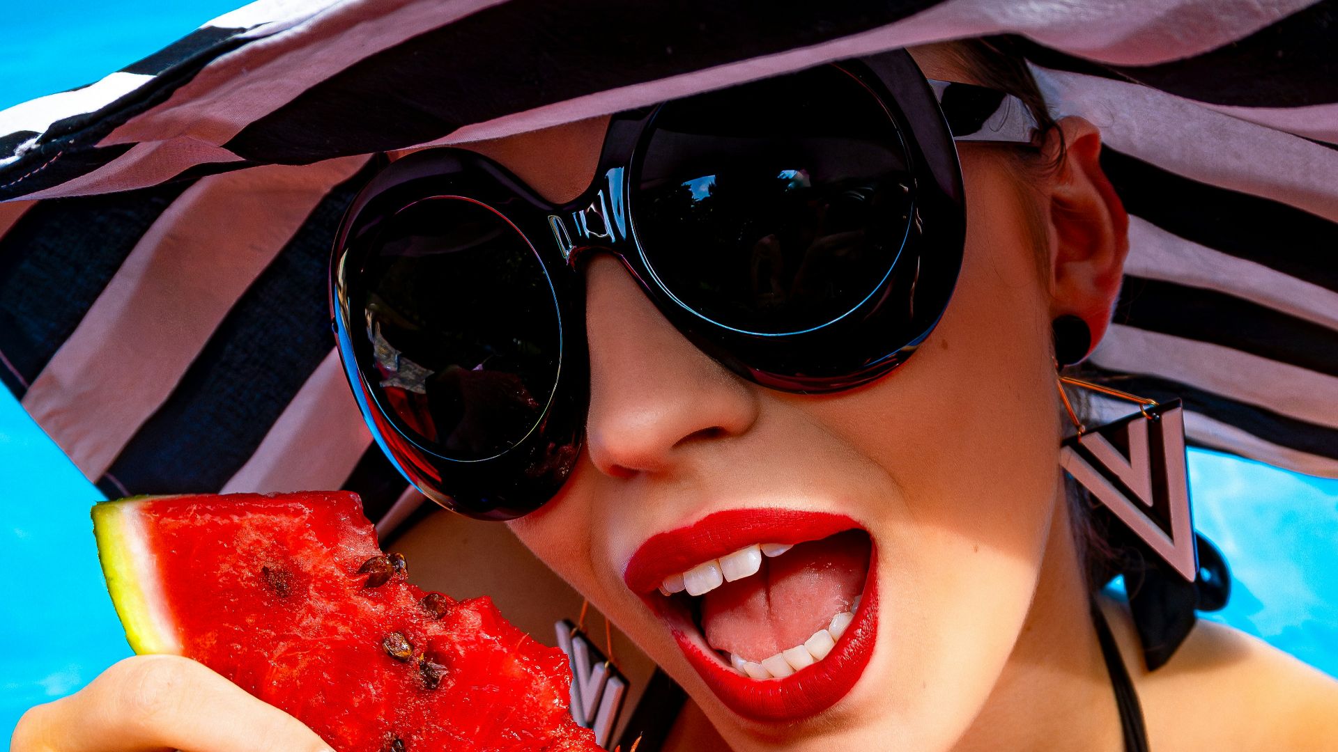 A woman wearing a hat and sunglasses holding a slice of watermelon
