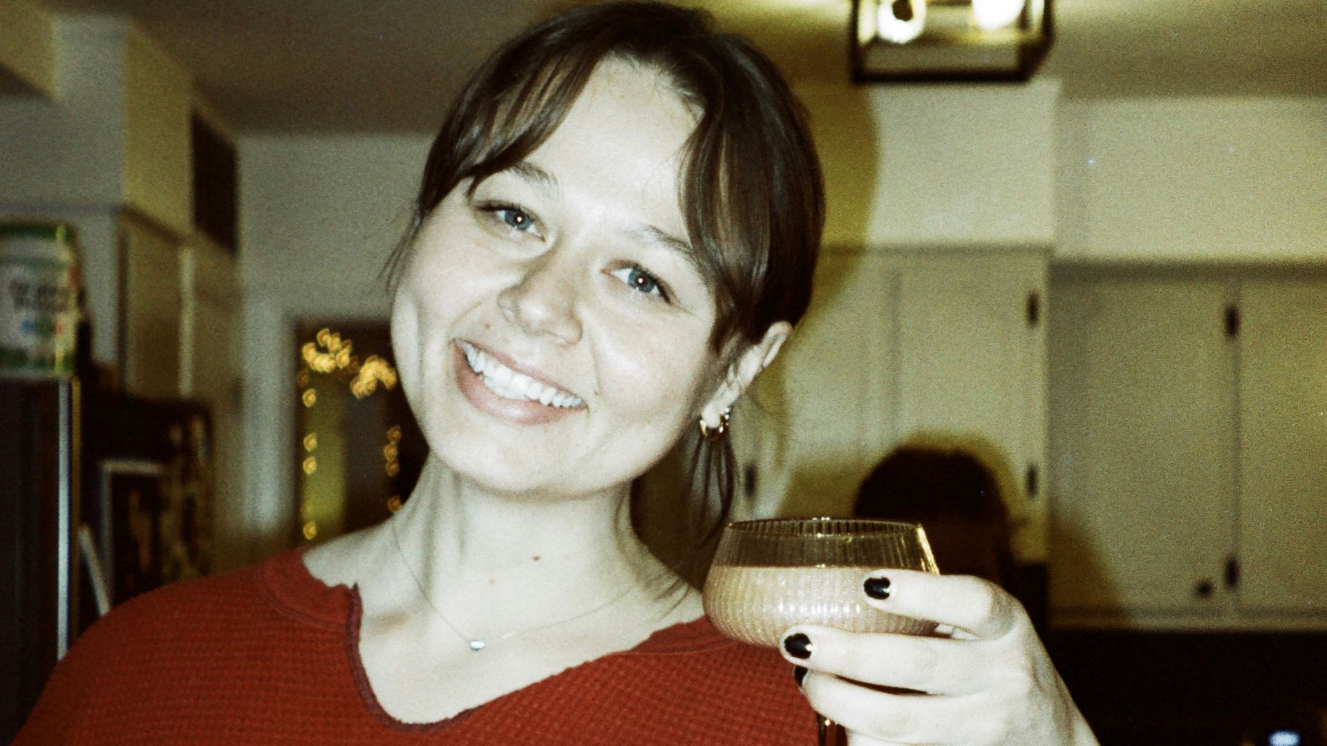 A woman smiles, holding a cocktail in her kitchen.