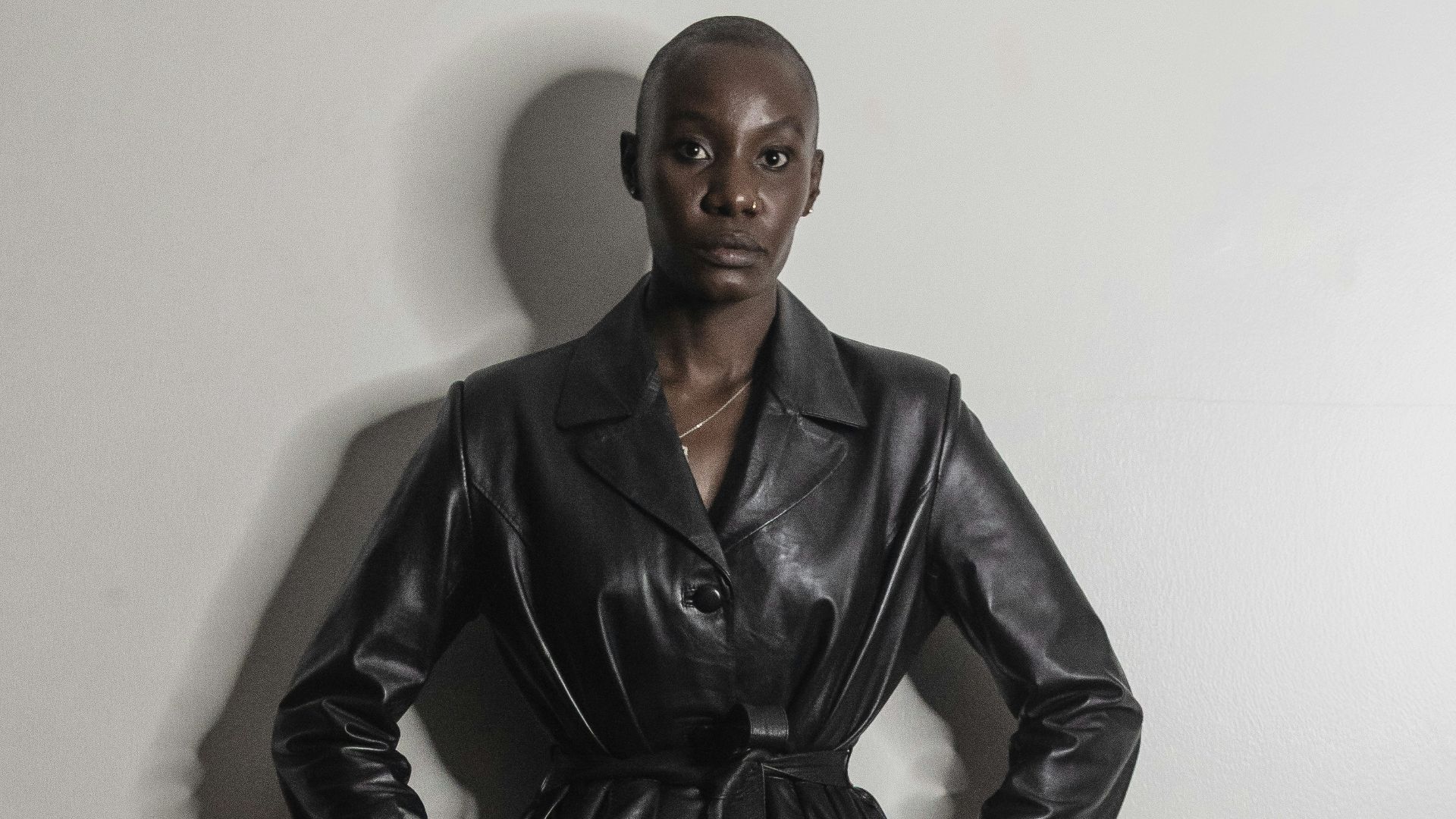 Woman in black leather coat and skirt poses against wall.