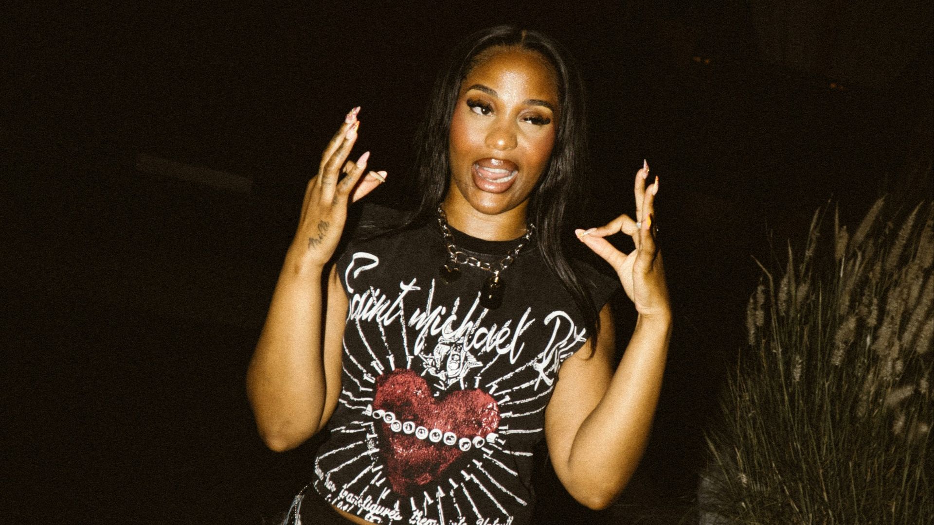 A woman poses with hands raised in a dark setting.