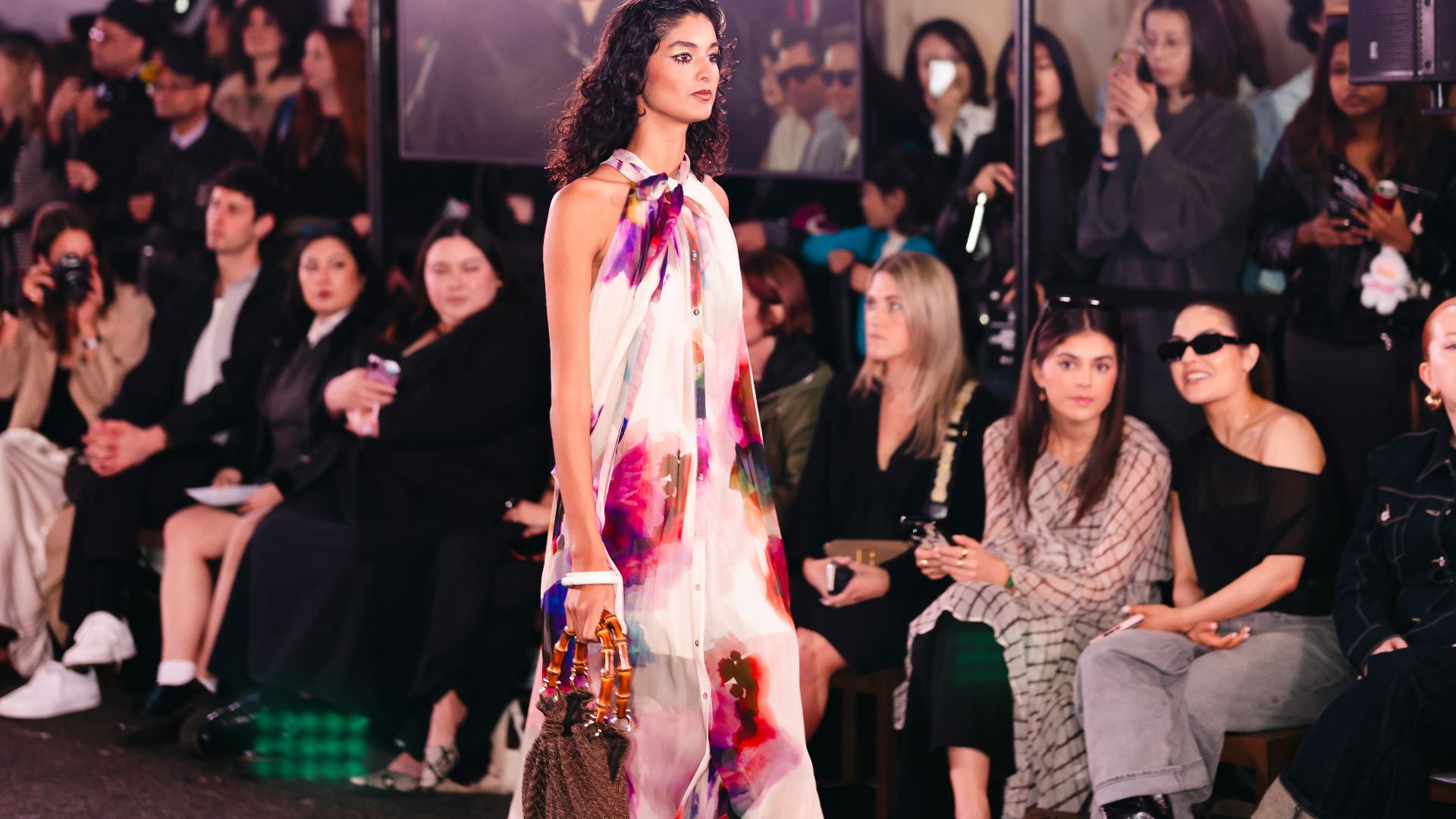 Model walks runway in floral dress with audience watching